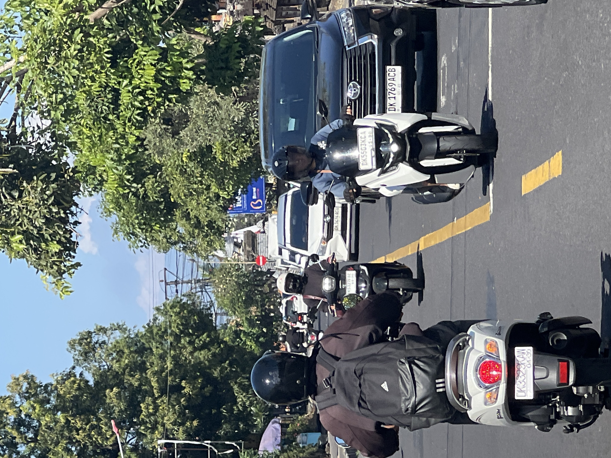 This is a vertical, eye-level shot of traffic on a sunny day in Sukawati, Indonesia. The scene is dominated by motorcycles and cars navigating a street lined with lush green trees and overhead power lines. In the foreground, two individuals are riding scooters. The scooter on the left, seen from behind, is silver and has a black backpack on the rider's back. The scooter on the right is white and black, with the rider wearing a light blue shirt and a dark helmet. A black SUV is visible behind the white scooter. Further down the road, other vehicles, including a white car, are present. The road surface is asphalt with yellow lane markings. The sky is clear blue with a few white clouds.