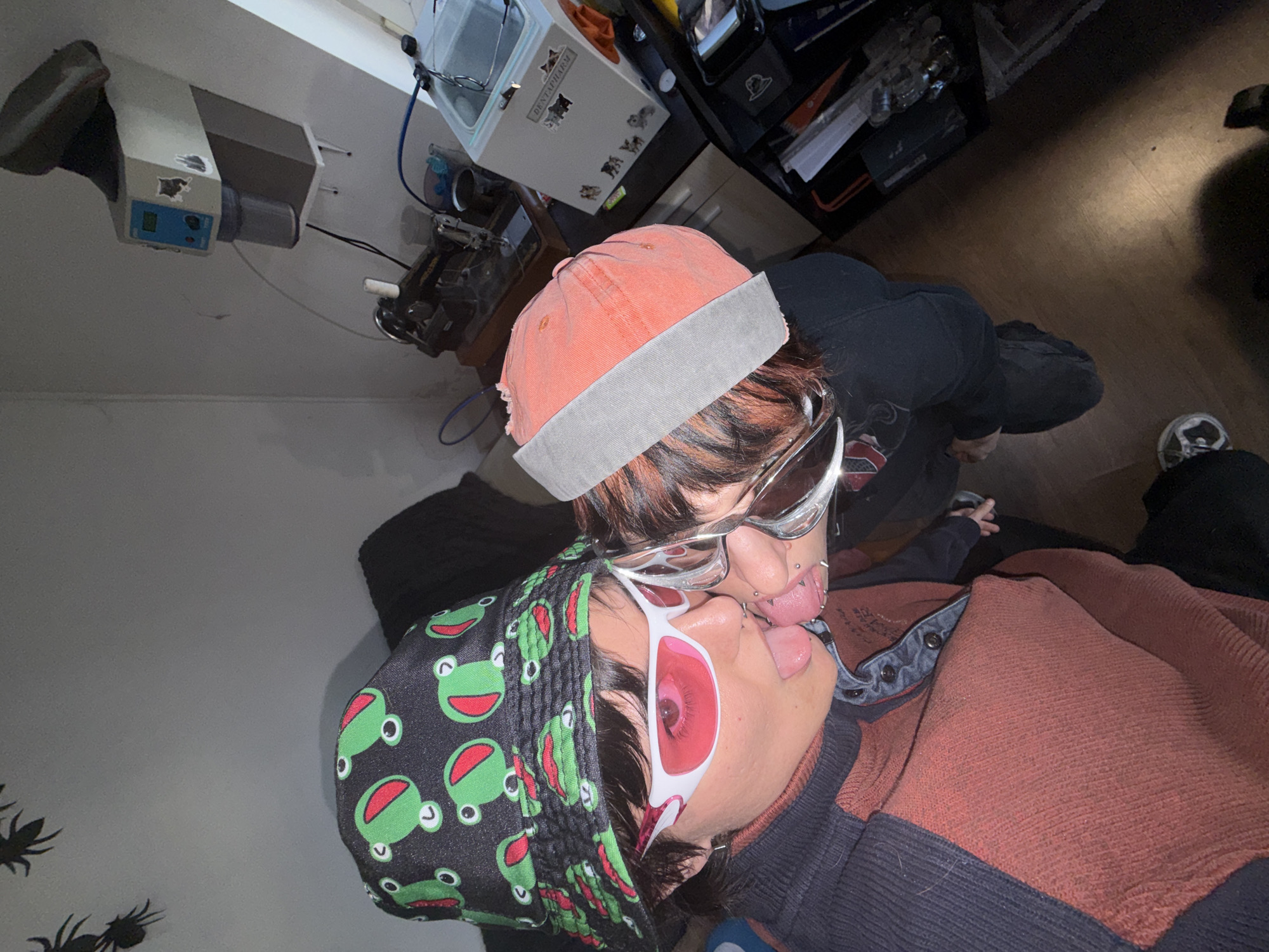 Two people are face-to-face, appearing to be kissing or close to kissing. The person on top is wearing a distressed orange and grey hat, silver sunglasses, black top. The person on the bottom is wearing a black head covering with green frog designs, red sunglasses with white frames. Both people have visible piercings. The room appears to be cluttered with objects, including some kind of medical device in the background, shelves with various items, and a floor that is partially hardwood. The location is unknown and could not be determined to be in Sofia, Bulgaria.