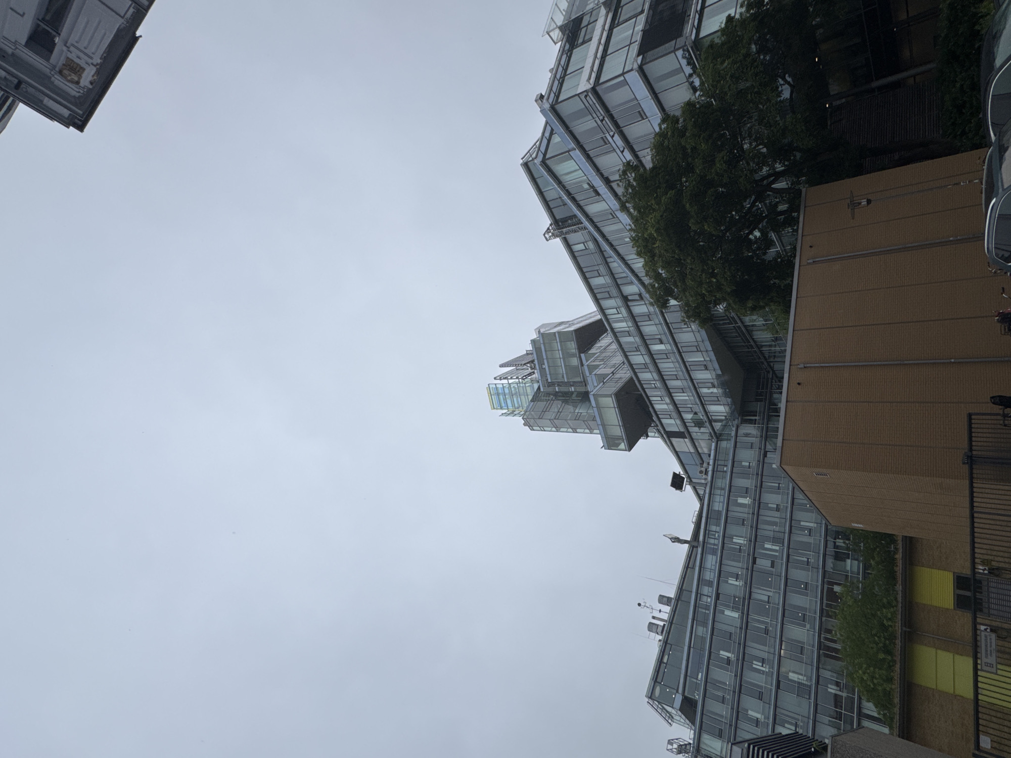 An exterior, low-angle, wide shot captures a modern, multi-story building with a striking, cantilevered glass structure at its apex. The building's facade is predominantly glass and steel, with a segmented, geometric design that gives it a dynamic, somewhat angular appearance. Trees and greenery are visible at the base of the building, with a brick structure also present in the foreground. The sky is overcast and a pale gray, contributing to a muted and somewhat diffused lighting condition across the scene. The location appears to be urban, with other buildings and some parked vehicles visible, though the specific city and surroundings are not identifiable from the image.
