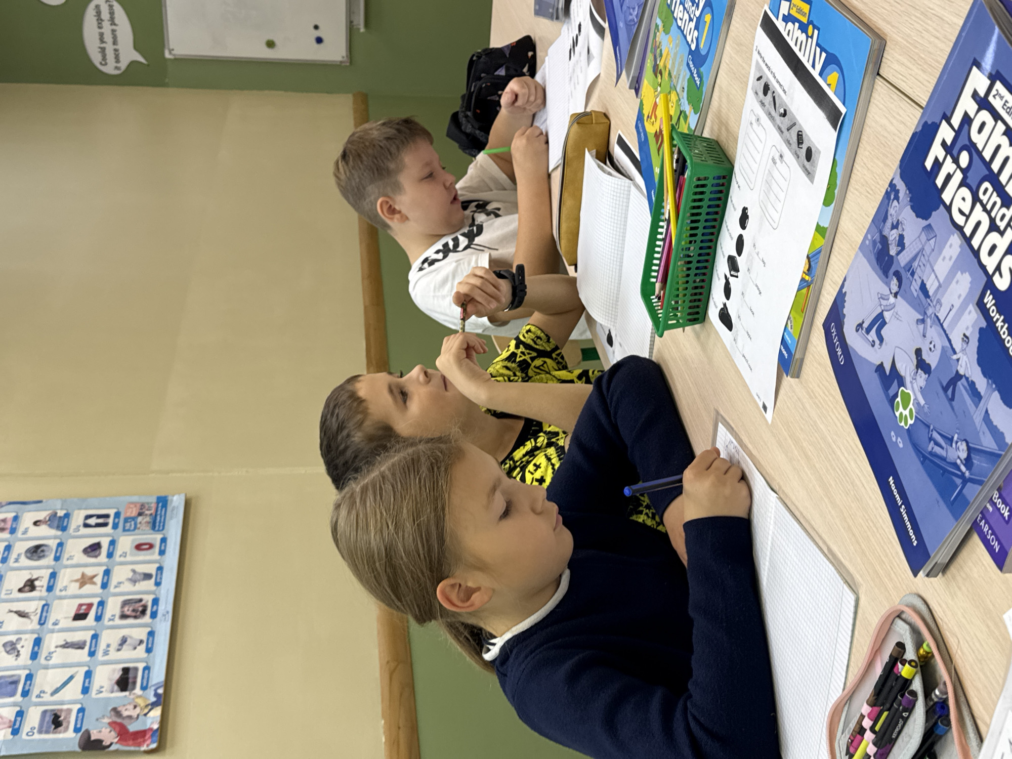A medium shot captures three children, two boys and a girl, sitting at a wooden table, engaged in classroom activities. The girl, in the foreground, wears a navy blue sweater with a white collar and is focused on writing in a notebook with a blue pen. A young boy, to her right, sports a black and yellow patterned shirt and is also writing, holding a pencil in his right hand. Another boy, further to the right, is seen from the side, wearing a white t-shirt with a black graphic print, and appears to be looking towards his notebook.
The table is littered with school supplies, including an open notebook with graph paper, a green pencil holder filled with colored pencils, a yellow pencil case, and several books. One book prominently displays the title "Family and Friends" with colorful illustrations. A partially visible alphabet chart hangs on the wall in the background, along with a whiteboard with a speech bubble containing the text "Could you explain it once more please?". The wall color appears to be a muted yellow-beige. The overall setting suggests a classroom or a study environment. The image does not provide any visual cues to determine its specific location within the city of Perm, Russia.