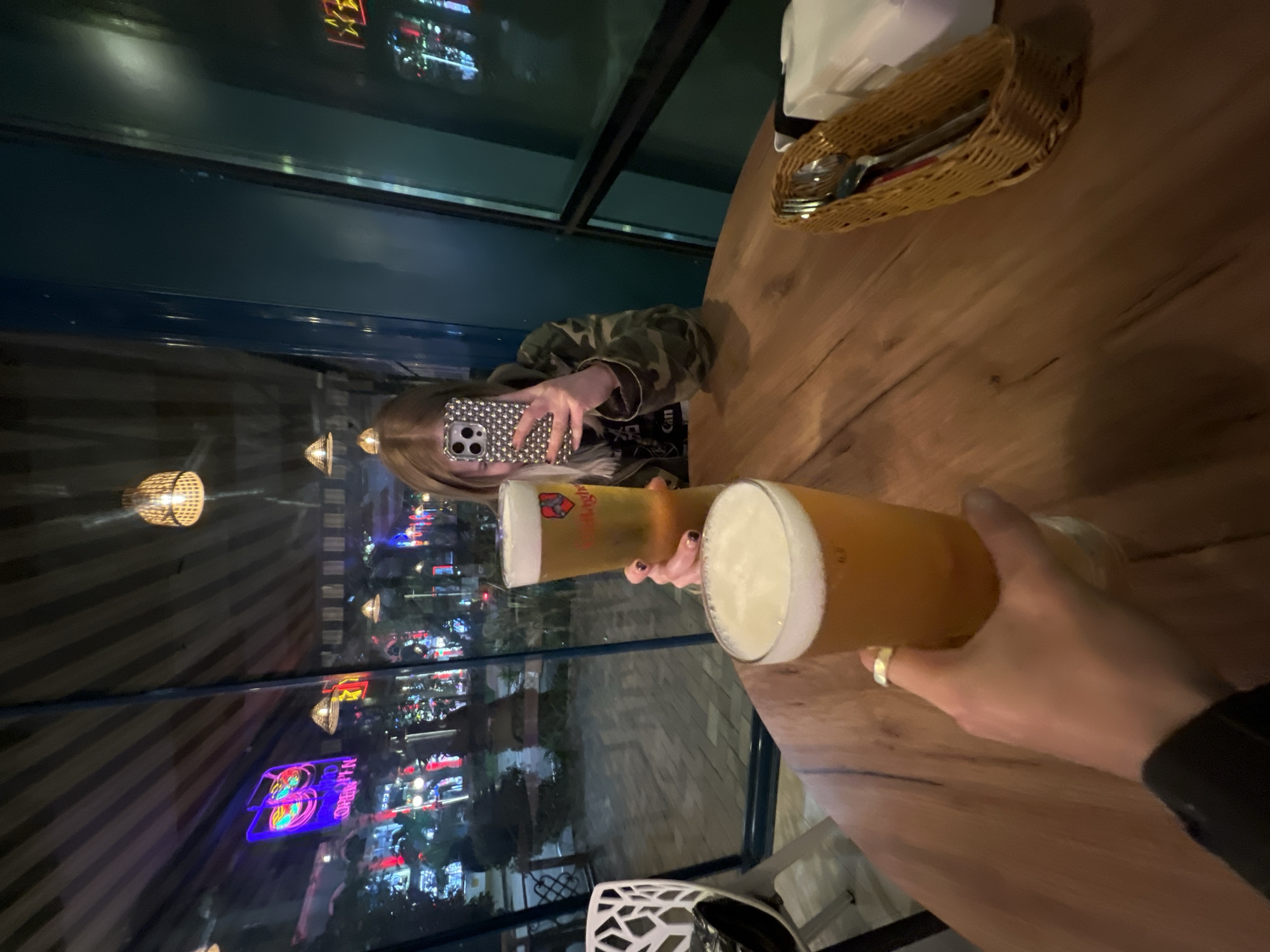 The image depicts a nighttime scene inside a restaurant, possibly in Sochi, Russia, as suggested by the reflection in the window of what appears to be illuminated buildings and signage in the Russian language. A person is holding up two tall beer glasses, likely in a toast or celebration. The beer appears to be a light-colored ale with a substantial head of foam. Another person, reflected in the window, is holding a smartphone, possibly taking a picture. The table is made of wood and there is a basket containing cutlery and napkins.