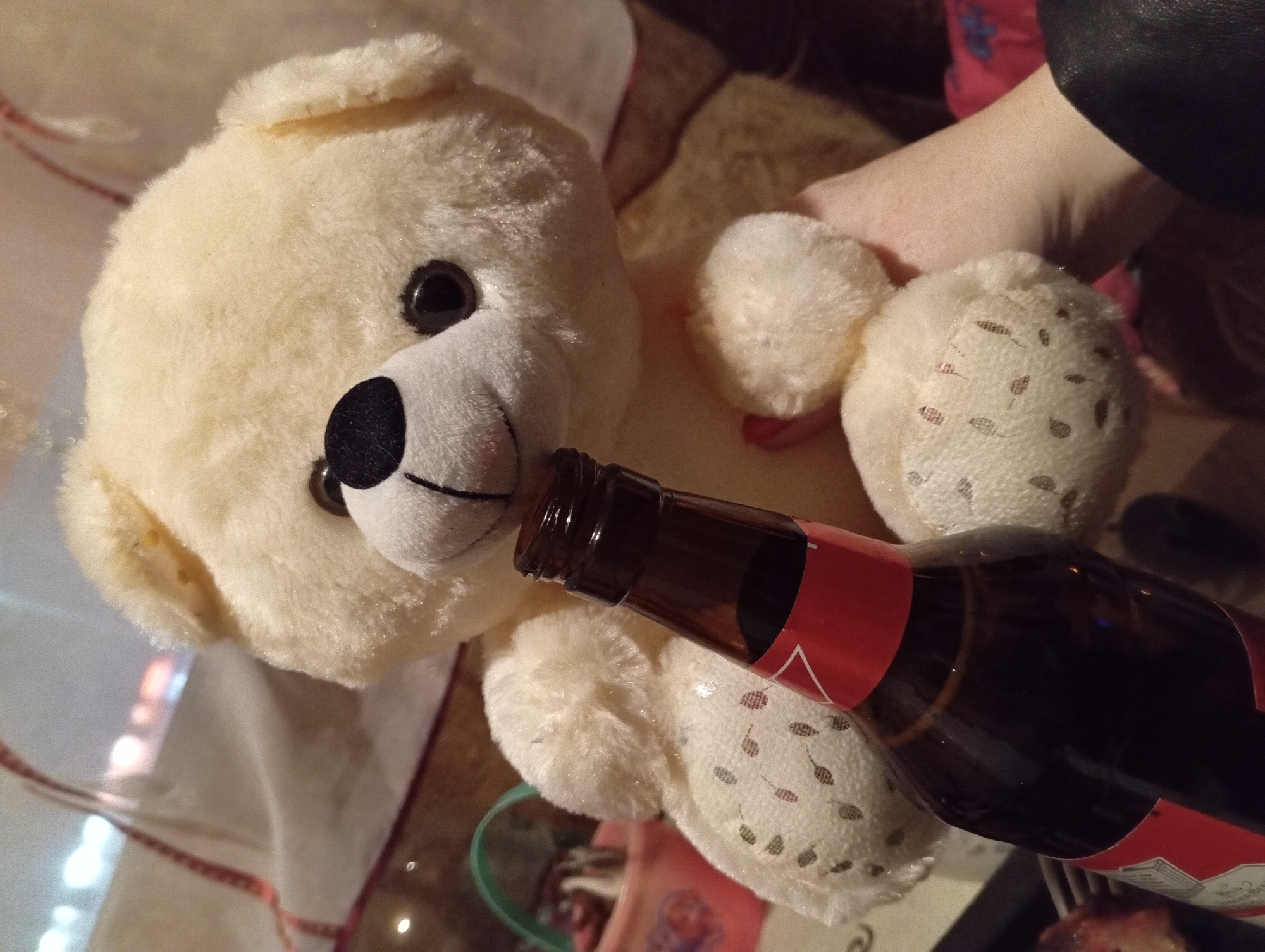 A light beige teddy bear with black eyes and a white muzzle is positioned horizontally on a table. A brown glass bottle with a red label is held by a person out of focus. The bottle's opening is near the bear's muzzle, as if the bear is drinking from it. The background shows glimpses of other items, including a cloth with a red edge, and what appears to be the edge of another table. The location and city are not evident in the image.