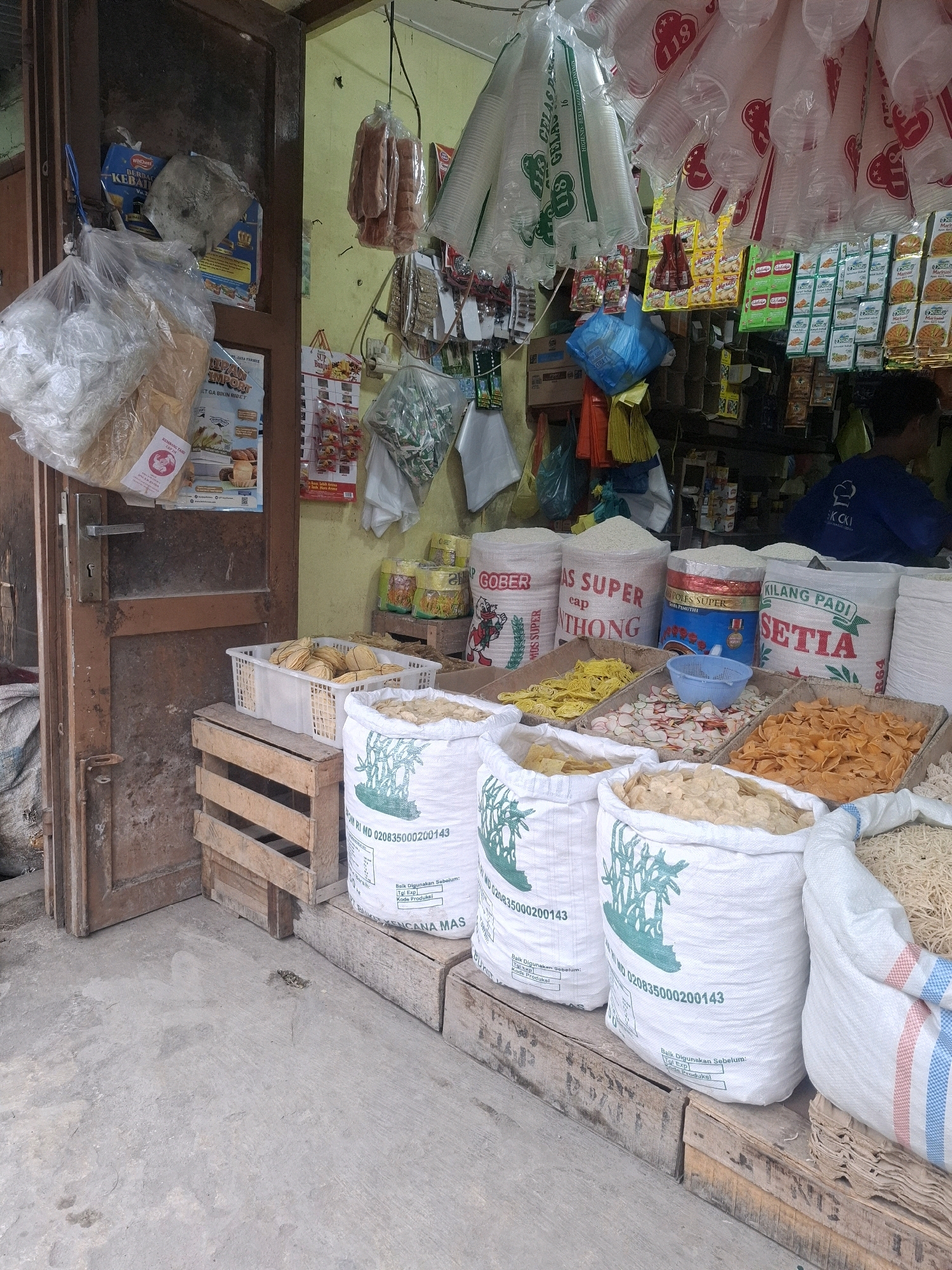 The image depicts an outdoor market stall in Kenangan, Indonesia. The stall is filled with various food products, including large sacks of rice or flour, baskets of snacks, and packaged goods hanging from the ceiling. In the foreground, several large white sacks with green illustrations of palm trees are visible, some containing dry goods like crackers or snacks. Next to them, wooden crates hold more baskets of food items. Behind these, various types of packaged snacks and seasonings are displayed. Hanging above the stall are numerous disposable cups and plastic bags filled with items. A person wearing a dark blue shirt is partially visible in the background on the right, appearing to be a vendor or customer. The stall is set up against a weathered wooden door and frame. The ground in front of the stall is made of concrete. The overall scene suggests a bustling local market environment.