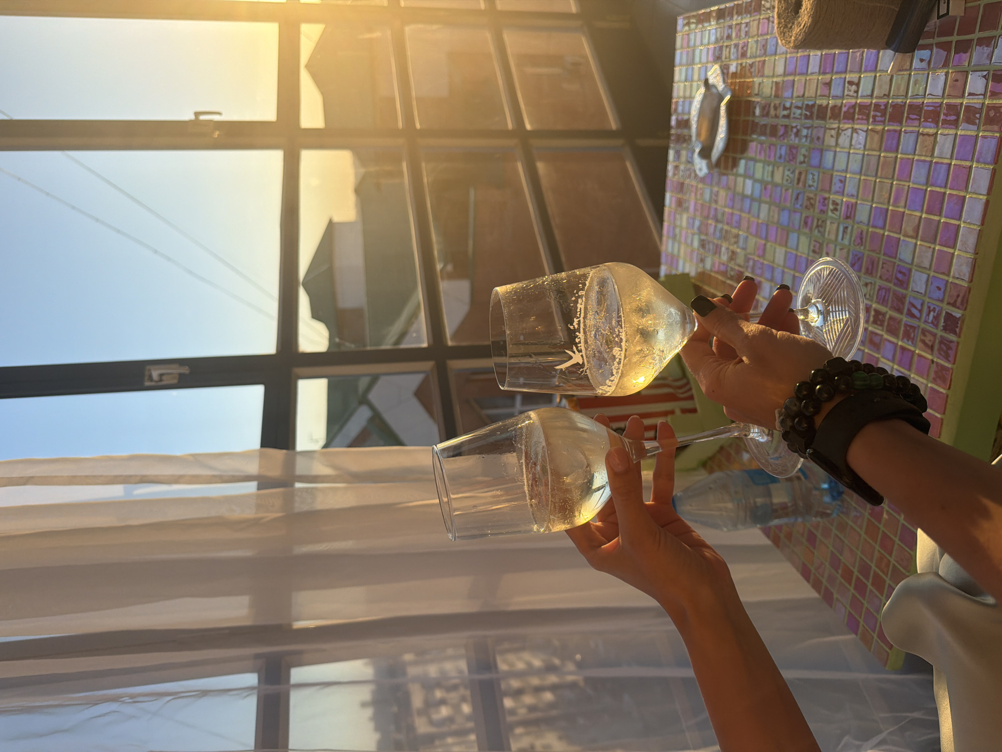 Two individuals perform a toast, clinking two stemmed glasses filled with effervescent pale yellow liquid in the city of Krasnoyarsk, Russia. The scene is set indoors by a large window overlooking a residential area at sunset.
The left hand, with light-colored fingernail polish, holds a glass aloft. The right hand, adorned with dark fingernail polish and a multi-beaded dark bracelet on the wrist, holds the second glass, which features a white goose logo near the rim. Both glasses contain visible bubbles.
In the foreground, a square table with a colorful mosaic tiled surface (composed of iridescent pink, purple, blue, green, and gold squares) is visible. On the table, from left to right, are a transparent plastic water bottle, a silver-colored fish-shaped ashtray, a spool of natural-colored twine, and a small dark rectangular object.
The background consists of a large, multi-paned dark-framed window. The left section of the window is partially obscured by sheer white curtains, which are illuminated by soft, indirect light. Through the window, the sky transitions from clear blue at the top left to a bright golden-orange hue on the right, indicative of sunset. Below the horizon, numerous residential buildings with varying roof designs (some green, some reddish-brown) are visible. The overall lighting is warm and golden, with strong sun glare emanating from the right side of the window.