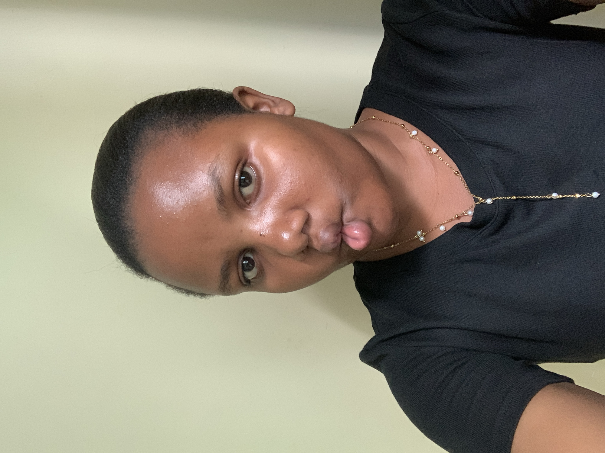 A young woman with dark hair pulled back is facing the camera with a playful expression, sticking out her tongue and pursing her lips. She wears a black crew-neck shirt and a delicate gold necklace with small white pearls. The background is a plain, light green wall. The location is indiscernible from this image and cannot be placed in Dar es-Salaam, Tanzania or any other specific location.