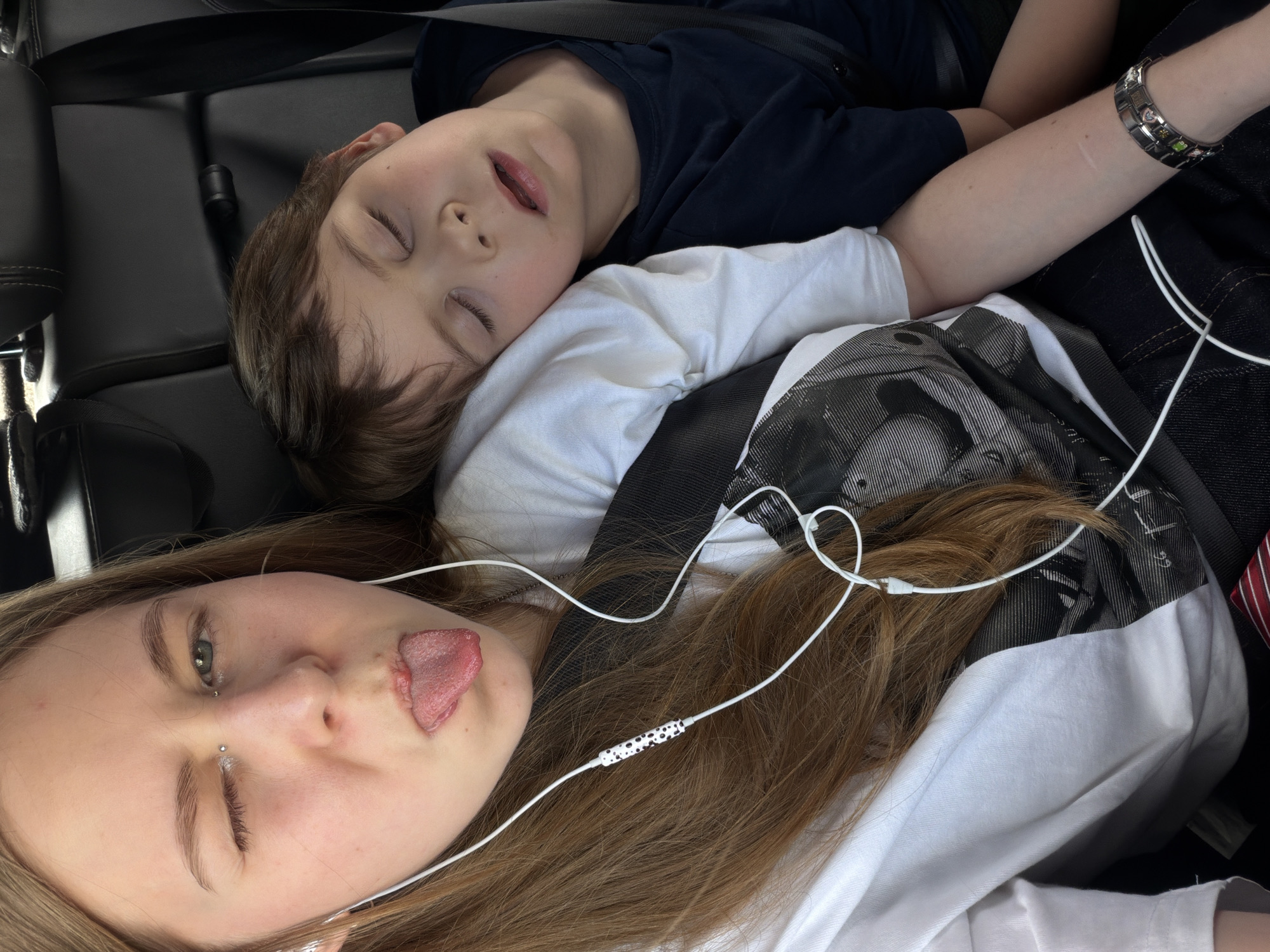 Two young people are in the back seat of a vehicle. A girl in the foreground, wearing a white t-shirt with a graphic print, is sticking out her tongue and winking. She has a piercing above her upper lip. White earbuds are in her ears, with the wire running across her chest. To her right, a younger boy is asleep, his head resting on her shoulder or on a pillow. He is wearing a dark blue t-shirt. The background consists of black leather car seats and a seatbelt. The scene is indoors, likely within a moving vehicle. The location is described as the city of Novoivanovskoe, Russia.