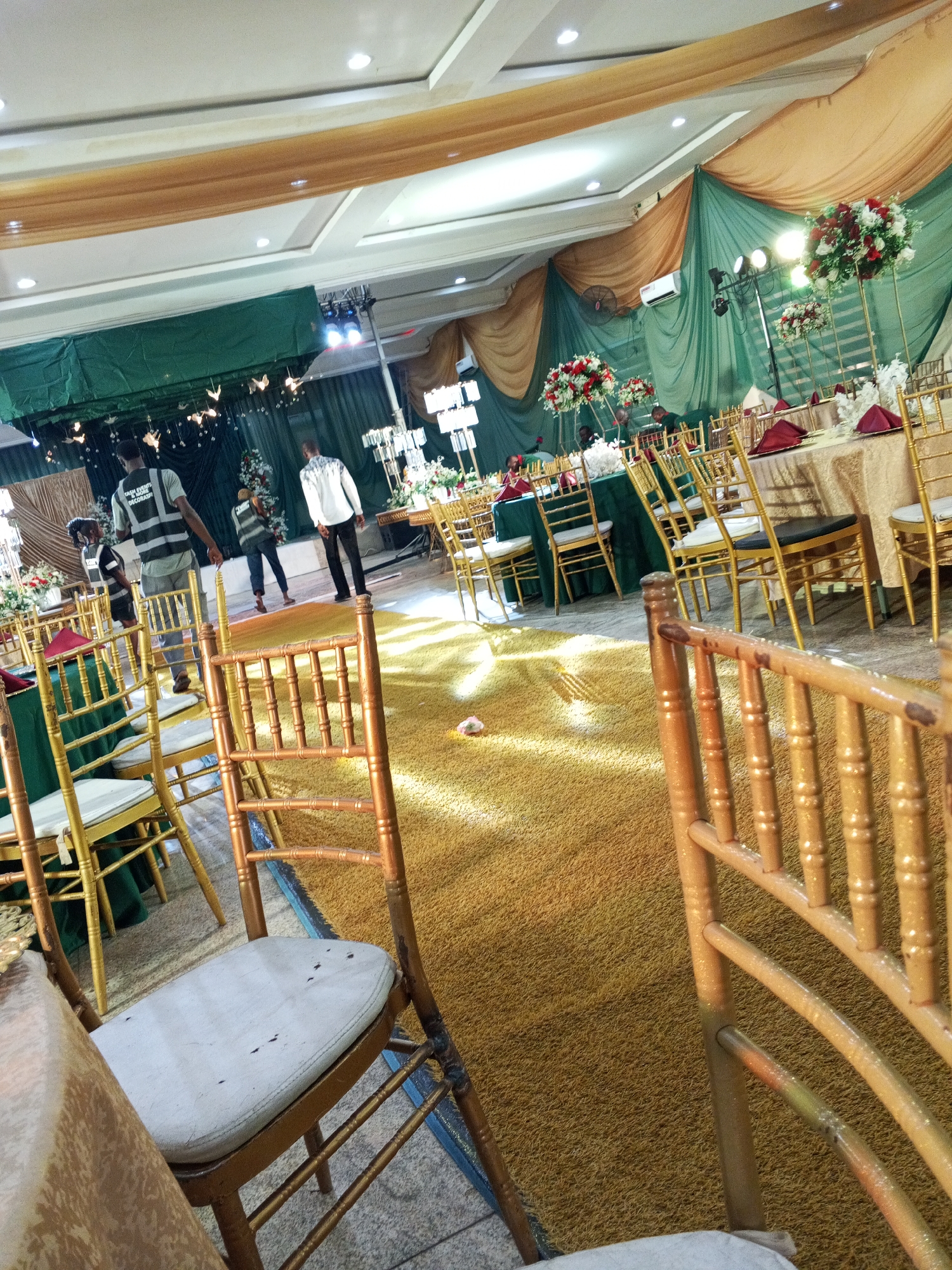 The image depicts an indoor event space, possibly a banquet hall, in Abuja, Nigeria. The hall is decorated with draped green and gold fabrics, and illuminated by overhead lights. Several round tables are set with beige tablecloths, maroon napkins, and floral centerpieces featuring red and white flowers. Gold-colored chairs are arranged around the tables.
In the background, a stage area is visible, draped in green fabric and adorned with lights and some decorative elements. A few individuals are present in the scene. Three men are walking across the venue. One man, wearing a grey vest with reflective stripes and the inscription "CHRIS EVENTS DECORATION," is walking away from the camera. Another man in a white shirt and patterned trousers is walking towards the stage. A third man, partially visible in the background, appears to be part of the event staff.
The floor is covered with a golden, textured carpet. The overall atmosphere suggests a prepared event, likely a wedding reception or a formal gathering. The presence of event staff and the table settings indicate an ongoing setup or preparation phase. The lighting suggests it is daytime, as natural light seems to be mixed with artificial illumination.