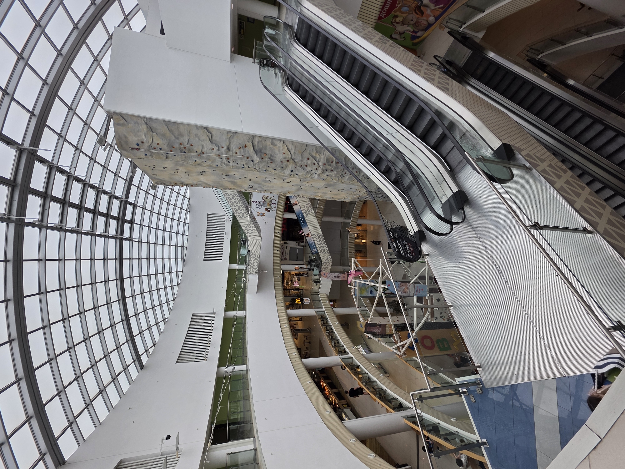 This wide-angle photograph, captured from a high vantage point within a modern multi-story shopping mall in Plovdiv, Bulgaria, looks down upon the lower levels and captures the expansive interior architecture. The image is significantly rotated clockwise.
On the left, a grand, curved glass and metal-framed structure forms a large wall or roof, allowing abundant natural light into the building. Beneath this, a prominent white architectural beam extends inward, from which a large rock-climbing wall is suspended, featuring numerous colored climbing holds.
To the right, multiple sets of escalators ascend, connecting the various floors of the mall. Below these, the mall's lower levels unfold, revealing a bustling commercial environment. Storefronts and restaurants line the curved walkways, with tables and chairs visible in dining areas. Geometric patterns adorn some wall sections and floor areas.
Several people can be seen moving about on the lower floors and near the escalators, engaging in typical mall activities. The overall lighting is bright, indicating daytime.
Visible text includes "JUMBO" on a colorful sign, likely indicating a children's store, "TELIOR" on a dark sign, and partially visible "PLAZA". Cyrillic text "ДОШЪЛ" (meaning "arrived" or "come") is also discernible on one of the upper level signs. Other indistinct signs are scattered across the shop facades. The scene conveys a sense of spaciousness, modernity, and active commerce within a vibrant public space.