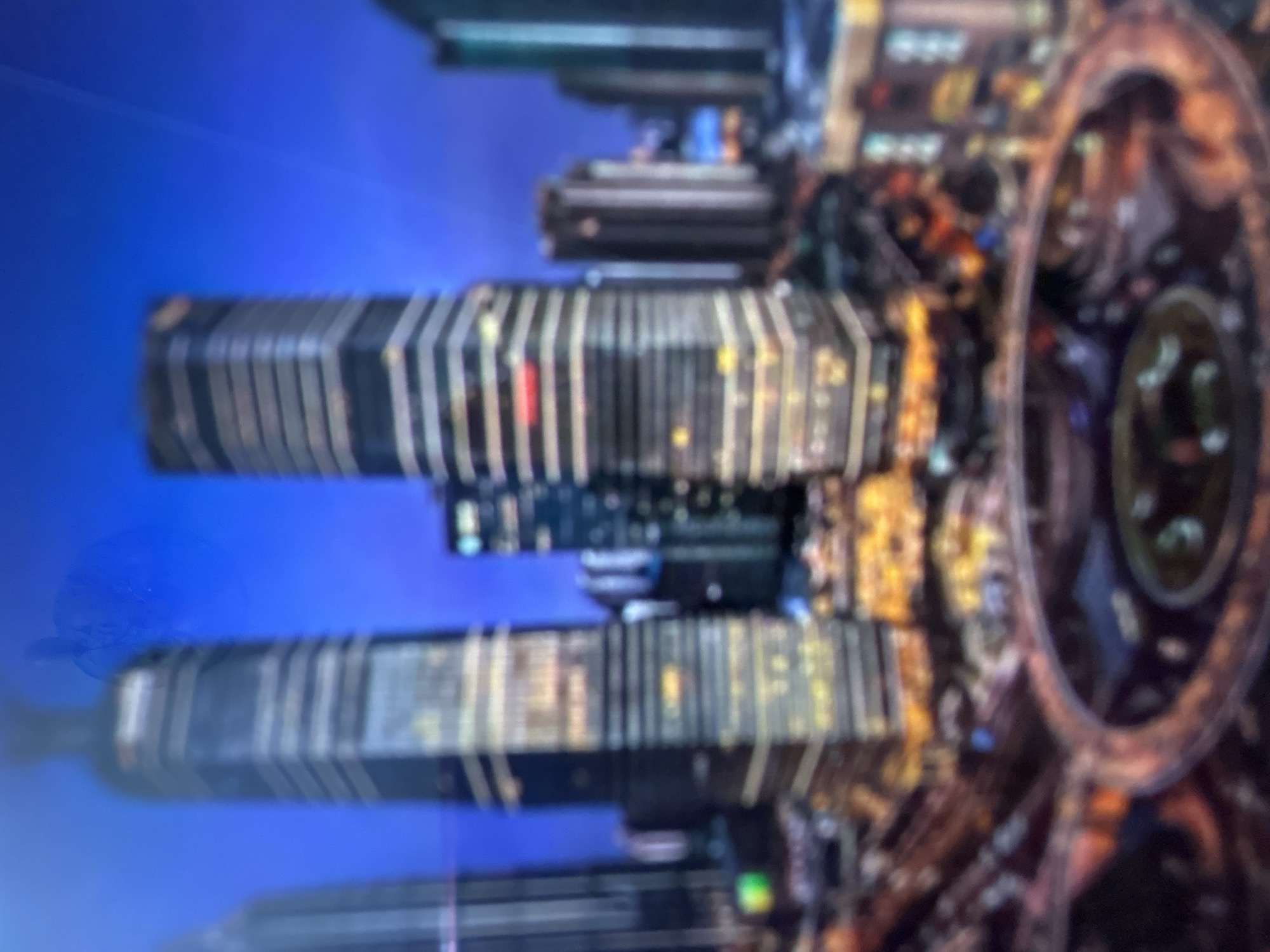 This media file shows a blurry, abstract image with a predominantly blue background. The main subjects are tall, rectangular structures with horizontal lines of light, suggesting buildings or perhaps a futuristic cityscape. These structures appear to be stacked or arranged vertically. There are also circular or ring-like elements visible on the right side of the image, also illuminated with lights. The lights within the structures and rings are yellow, white, and some red, indicating artificial illumination. The overall impression is one of a nighttime scene or an interior with dynamic lighting. The blurriness of the image prevents any specific identification of people or detailed activity. There are no discernible text elements. The image has a somewhat digital or computer-generated aesthetic due to its abstract nature and the clarity of the blue background.