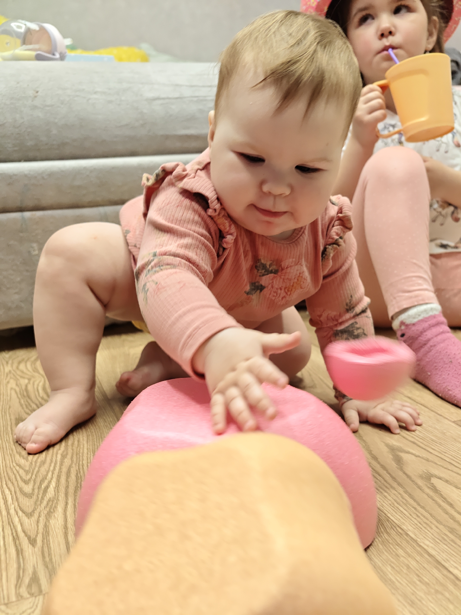 The image depicts a young child, likely a baby, crawling on a wooden floor. The child is wearing a pink long-sleeved top with a floral design. Another child, who is older, sits behind the crawling child, drinking from an orange cup using a straw. The older child wears a pink hat and pink leggings. A portion of a grey sofa is visible in the background. Toys can be seen in the background. There is no discernible evidence to indicate that the location is the city of Chaikovsky, Russia. The primary action in the image is the younger child reaching out towards a pink and tan toy.