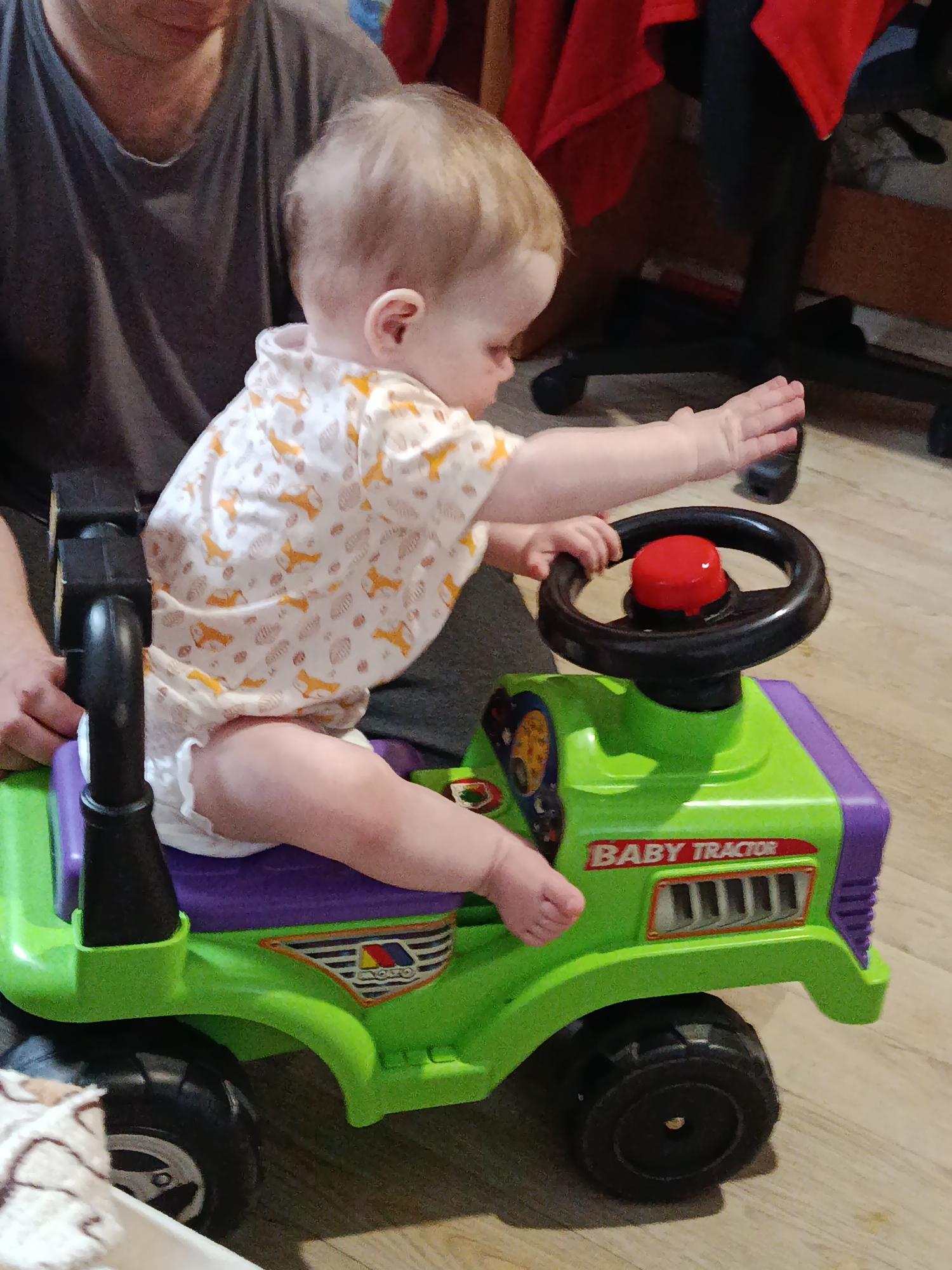 An indoor photograph taken in Фоки, Russia, captures a light-haired baby sitting on a green and purple "BABY TRACTOR" ride-on toy. The baby, wearing a white romper with an orange fox and leaf pattern, and a diaper, has their left arm extended forward in a gesture of play, while their right hand rests on the toy's black steering wheel. A prominent red button is visible on the steering wheel.
A partially visible adult, wearing a grey t-shirt, is seated behind the baby, appearing to support or supervise. The "BABY TRACTOR" toy features a vibrant green body with purple accents on the seat and front grill. The text "BABY TRACTOR" is clearly visible on the side, along with a circular logo that includes the word "KYOBO" beneath a red, blue, and yellow striped design. The toy is equipped with chunky black wheels.
The setting appears to be a home, indicated by the light wooden floor, casual attire, and a glimpse of a black office chair and a red draped cloth in the background. The baby seems engaged and focused on playing with the toy. The indoor lighting suggests either daytime or well-lit artificial illumination.