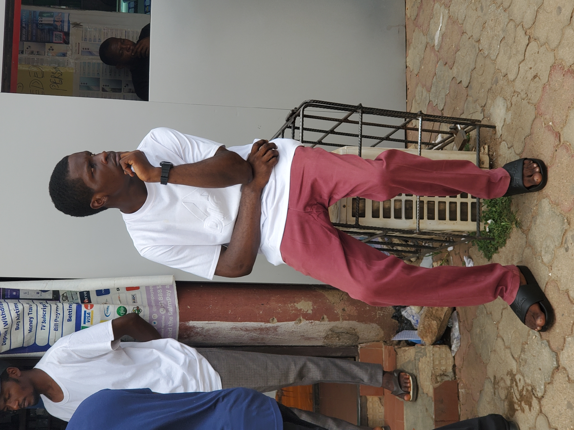 The image depicts a man standing outside a shop in Jos, Nigeria. He is wearing a white t-shirt, maroon pants, and black sandals. He has a black watch on his left wrist and is resting his chin on his hand, appearing thoughtful. In the reflection of the shop window behind him, another man is visible. Below him, partially obscured, are two other people wearing white t-shirts. A sign advertising financial services, including money transfers and bill payments, is visible on the building behind the man, with the logos of Visa, Mastercard, and Verve visible. The ground is paved with a patterned brick. There is a metal grate next to the man.