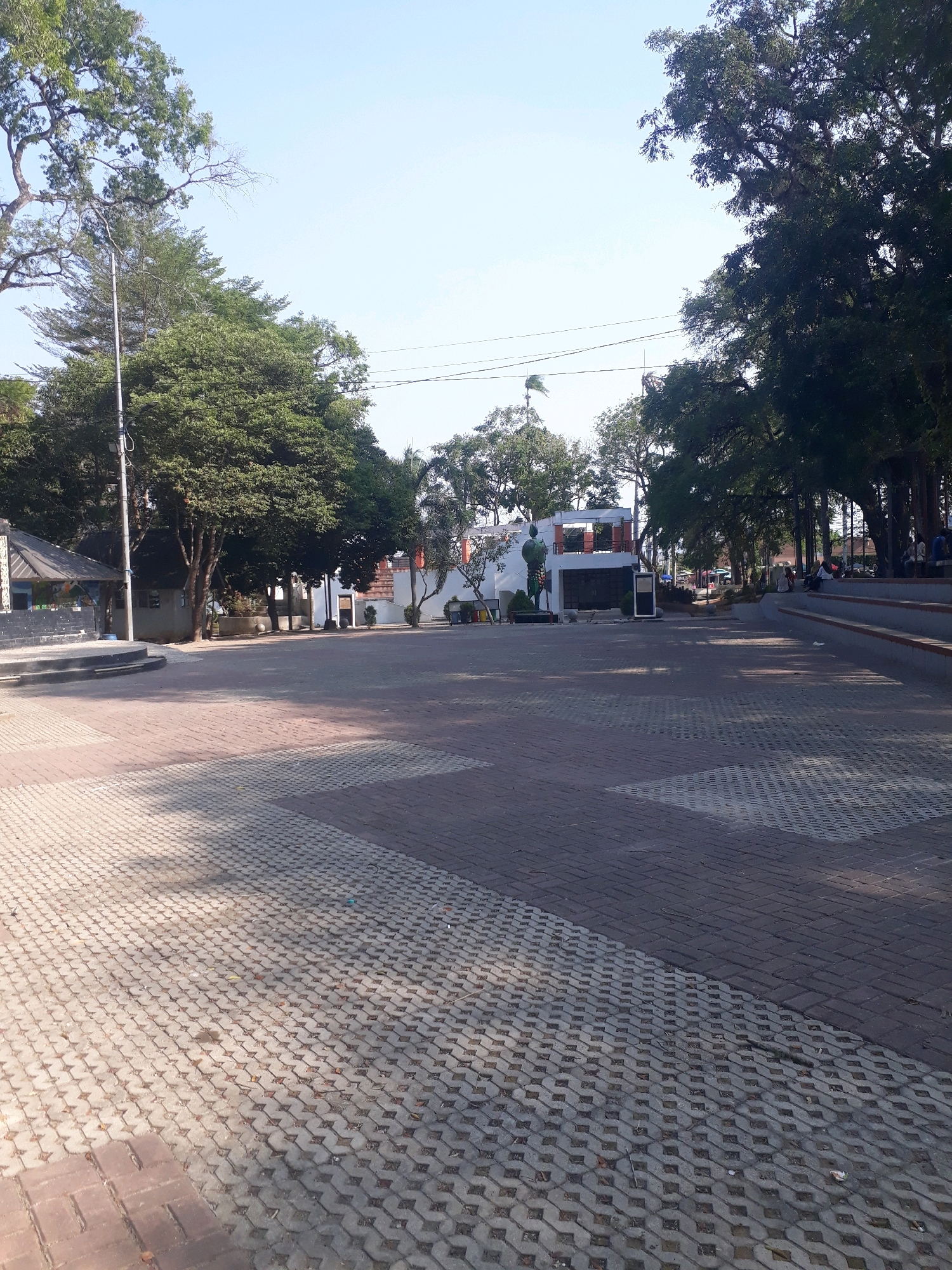The image captures a view of the Alun-Alun Taman Merdeka in Pangkalpinang, Indonesia. The foreground is dominated by a paved area, with two distinct types of interlocking concrete pavers. The left side features a lighter-colored, more open-patterned paver, while the right side uses a darker, denser pattern. Sunlight casts shadows across the paving.
In the mid-ground, a slightly elevated area with tiered seating is visible on the right, with several individuals seated. A walkway or path leads into the distance. On the left, a small, open-sided structure with a dark roof and visible support beams is present, along with a stage-like platform. Further back, a modern building with white walls, a flat roof, and dark window frames stands. This building appears to be part of the park's amenities.
The background is filled with lush green trees, creating a canopy over parts of the park. A clear, light blue sky stretches across the top of the image, suggesting a bright, sunny day. The overall impression is of a public park space, likely used for recreation and community gatherings.