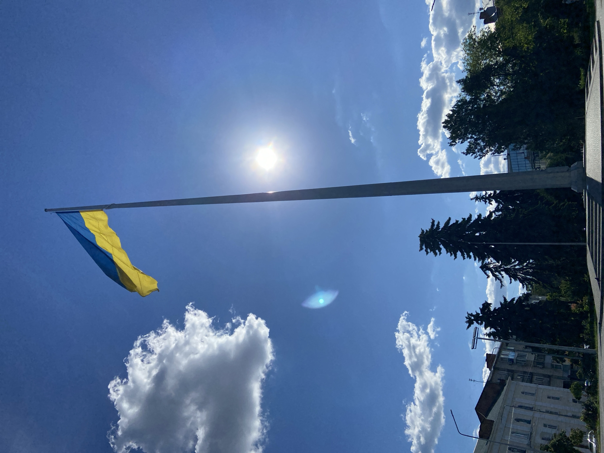 A Ukrainian flag flies from a tall flagpole against a bright blue sky on a sunny day. The flag, with its horizontal stripes of blue and yellow, is partially illuminated by the sun, which appears as a bright, lens-flared orb in the upper portion of the frame. Scattered white clouds drift across the sky, adding texture to the scene.
In the background, the tops of dark green coniferous trees are visible, along with some of the upper floors of buildings. A hint of an urban setting is suggested by the presence of architecture and what appears to be a sidewalk or paved area in the lower right corner. The perspective is from a low angle, looking upwards towards the sky and the flag. The sun's intense light creates a lens flare, a circular artifact of light, in the middle of the sky.
The overall atmosphere is one of brightness and openness, with the flag symbolizing national pride against a backdrop of clear weather. The time of day appears to be midday due to the high sun and bright illumination. No people are visible in the image, and no text is discernible. The context of Poltava, Ukraine, suggests this is a public display of the national flag.
