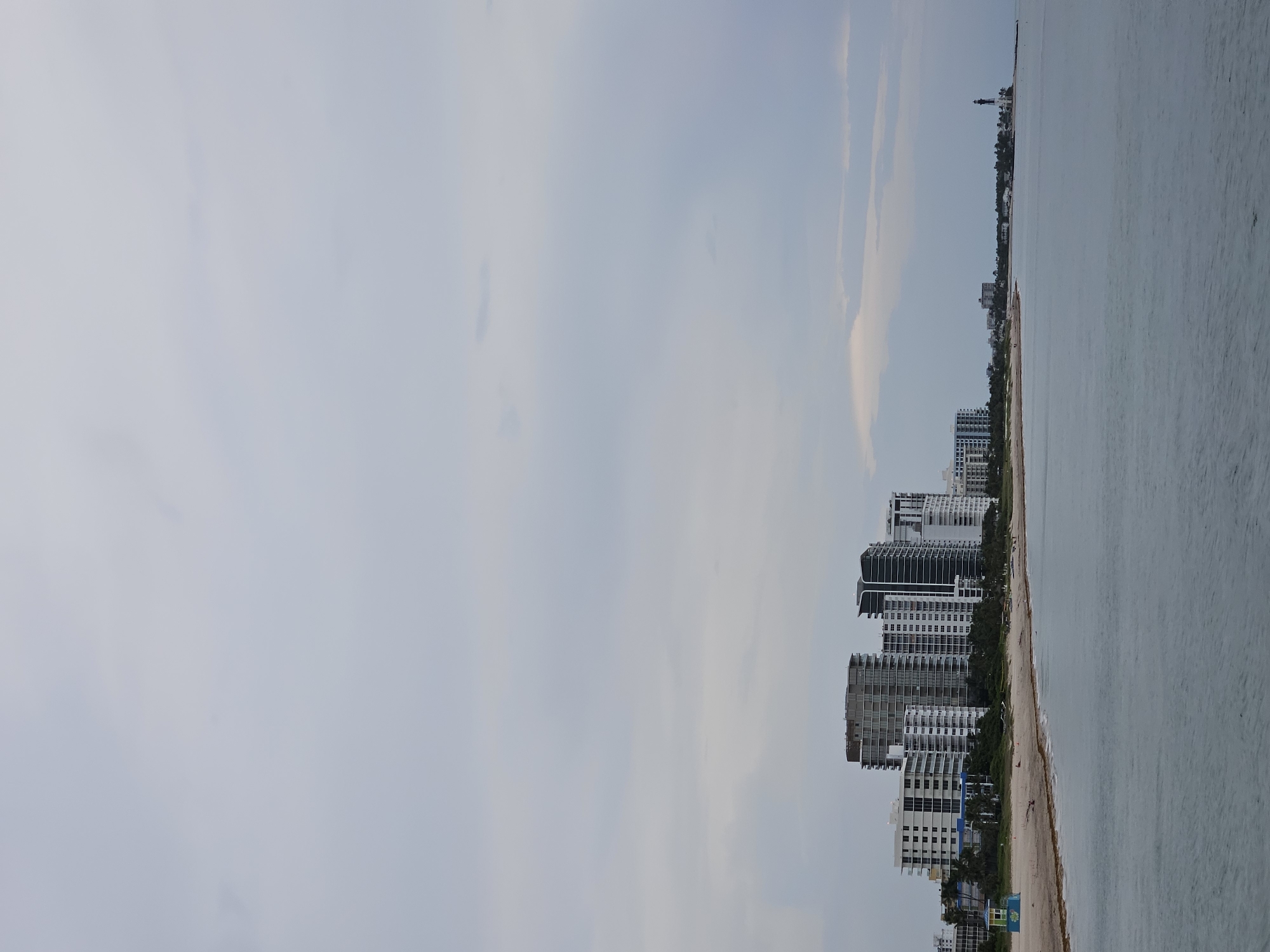 The image shows a view of the Pompano Beach coastline in Pompano Beach, Florida, USA. The scene is captured on an overcast day with a cloudy sky dominating the upper portion of the frame. In the mid-ground, a line of modern high-rise buildings stands against the horizon. These buildings are a mix of white and darker facades, with many featuring balconies. A sandy beach stretches along the foreground, meeting the calm, gray ocean. To the left, a small, brightly colored lifeguard station is visible on the beach. Further to the right, along the coastline, a lighthouse can be seen, indicating a more developed or historical area. The water is textured with gentle ripples. There are no discernible individuals engaged in specific actions within the frame, only the presence of the built environment and the natural elements of the sea and sky.