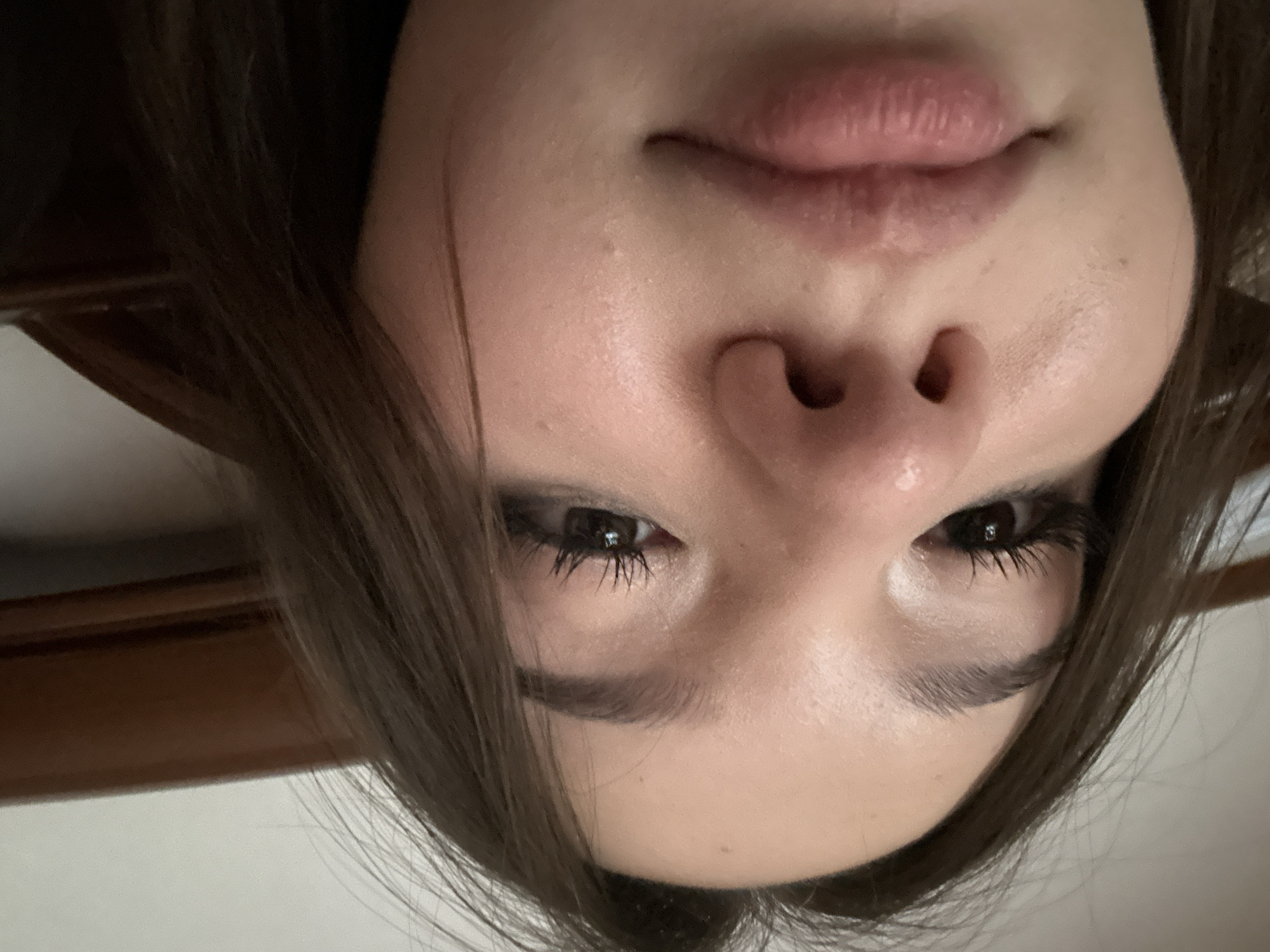 This close-up portrait features the upper half of a person's face, presented in an inverted orientation. The subject, likely a young woman, has her nose and mouth positioned towards the top of the frame, while her eyes and forehead are towards the bottom.
Her features include full, naturally pink lips that are slightly parted, and a prominent nose. Her dark brown eyes are open and gaze directly, with long, dark eyelashes, possibly enhanced with mascara or extensions. Her eyebrows are dark and well-defined. Dark brown hair frames her face on both sides. The skin appears fair, exhibiting a slightly dewy or oily sheen, particularly noticeable around the nose and cheeks, where some minor skin imperfections are visible.
The background is out of focus, suggesting an indoor setting. Hints of a light-colored wall and a wooden architectural element or furniture can be seen. The lighting is soft and diffused. The subject's expression is neutral, primarily focused on the camera. No text is visible in the image. The photo was taken in Naberezhnye Chelny, Russia.