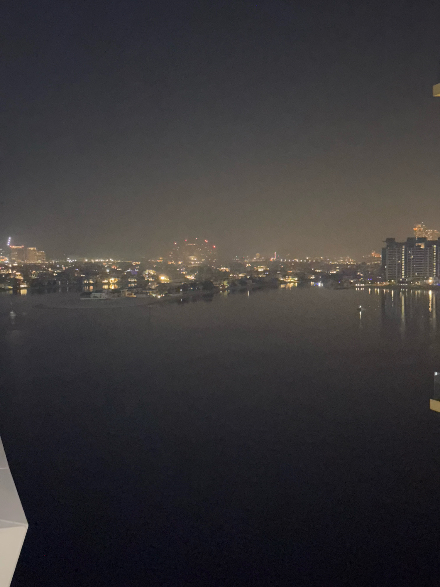 This image shows a nighttime view of the Dubai skyline, with the Dubai Marina area prominently visible. The foreground is dominated by dark water, likely a canal or the sea, reflecting the city lights. In the middle ground, a low-rise cityscape with illuminated buildings and houses lines the waterfront. Beyond this, taller apartment buildings and skyscrapers with rows of lights pierce the hazy night sky. The overall atmosphere is serene and somewhat muted due to the nighttime conditions and possible mist or fog. No people or specific actions are discernible in this wide-angle shot.