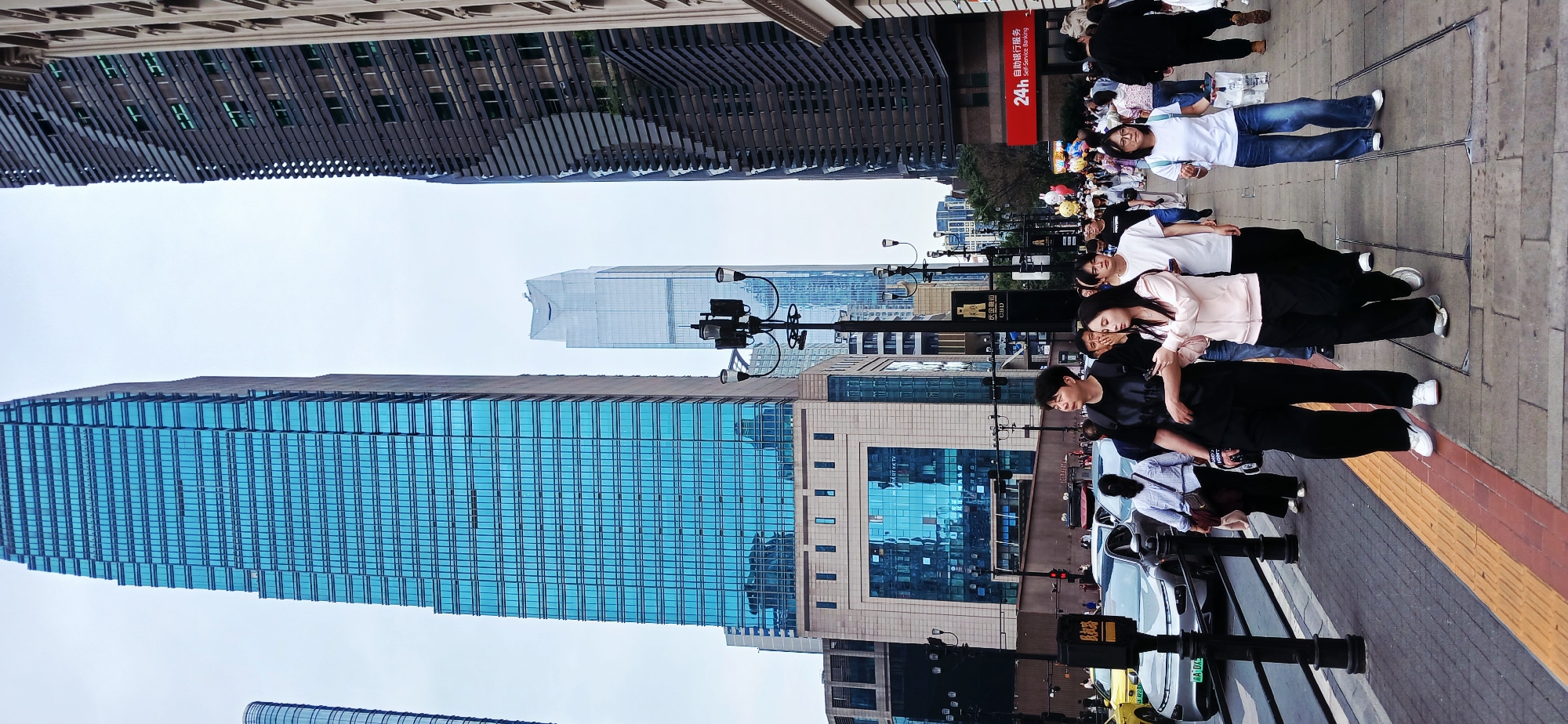 The image presents an urban scene, evidently in a city with modern architecture, featuring a group of people lying down on a sidewalk. The perspective is slightly tilted.
Several high-rise buildings dominate the background. One is characterized by a blue glass façade, while others display varying architectural styles, including one with a dark, linear pattern. The sky is overcast.
In the foreground, a line of individuals is lying horizontally along the sidewalk. Most are young, dressed in casual clothing such as jeans, t-shirts, and sneakers. Some appear to be interacting with their phones or resting. The sidewalk is paved and includes a yellow tactile paving strip.
Various street furniture and signage are visible, including street lamps, signposts, and a sign written in Chinese characters with "24h Self-Service Banking" written below in English. The sign also says, in Chinese, "Automatic self-service banking." The sign is red with white letters. In another sign, one can decipher "CHD" and "CBD." Some vehicles, including taxis, are partially visible along the street.
Based on the signage and architectural style, and the OCR text mentioning "Minquan Road," the location is likely within the Yuzhong District in Chongqing, China.