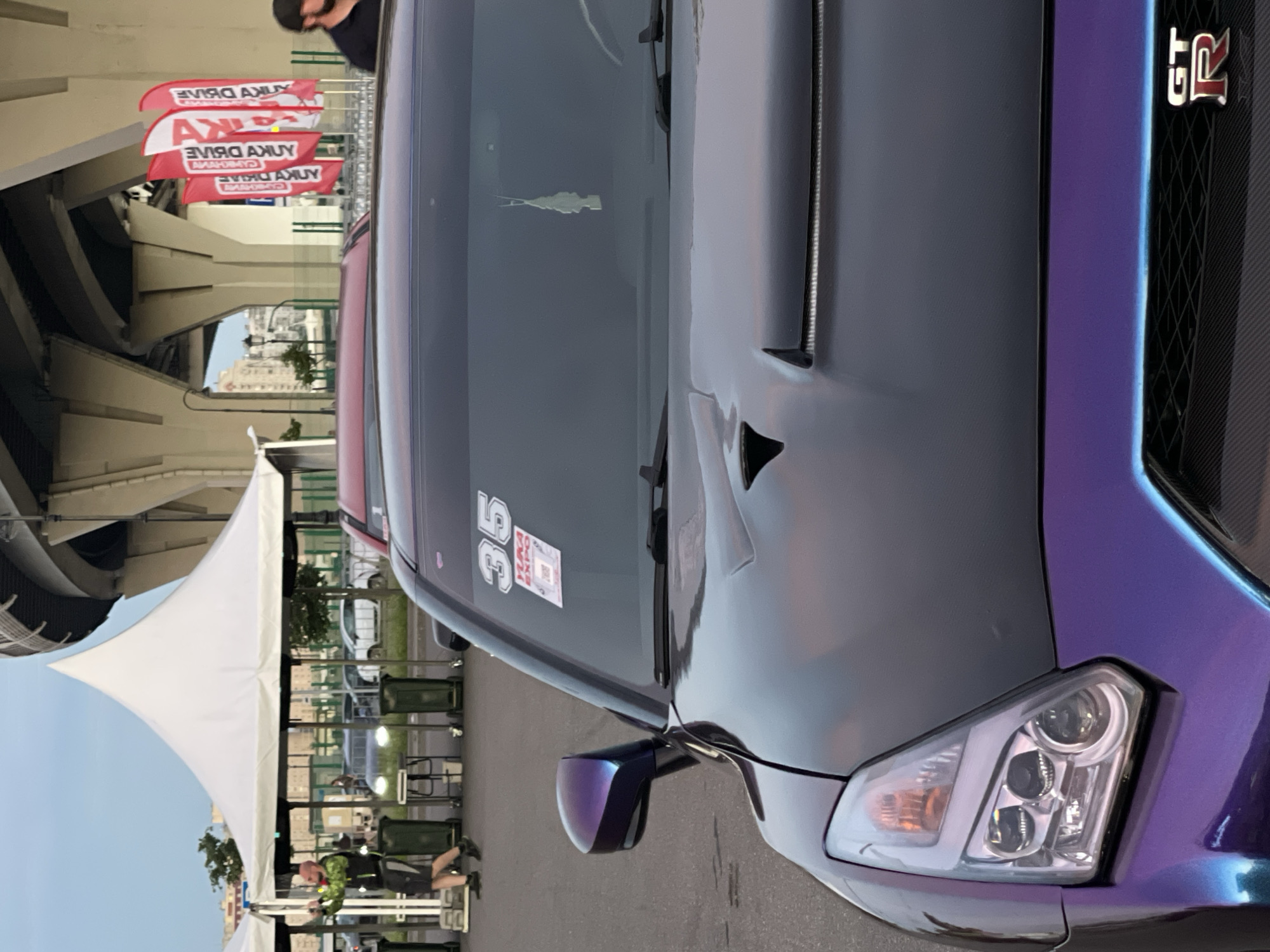 A wide shot captures a purple and black Nissan GT-R sports car parked on asphalt, with its front end prominently displayed. The car features a unique iridescent paint job and a carbon fiber grille with a red "GT-R" emblem. A white sticker with the number "35" and a QR code is visible on the windshield, along with a small, white, tree-shaped air freshener hanging from the rearview mirror. In the background, a white tent, a man in a green vest and black shorts, and several red banners with white lettering are present. The scene appears to be outdoors, possibly at a car show or event, with a large, concrete structure towering over the background.