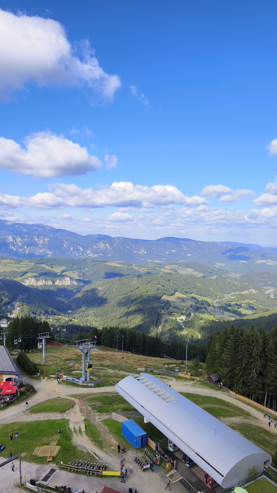 The image is a high-angle shot of a ski resort area in Semmering, Austria, on a sunny day with scattered clouds. In the foreground, a modern, curved, silver-roofed building appears to be a ski lift station, with several ski gondolas visible inside and on the tracks. To the left of this building, a smaller structure with a red awning and outdoor seating is visible, likely a restaurant or cafe. A ski lift with multiple chairs and towers extends into the background, crossing an open grassy slope. Further back, a winding road is visible, along with more ski lift infrastructure and trees. The background features rolling green hills and mountains, characteristic of the Austrian Alps. Several people are present in the scene, walking around the buildings and ski lift area. The overall atmosphere is one of a recreational area in a mountainous landscape.