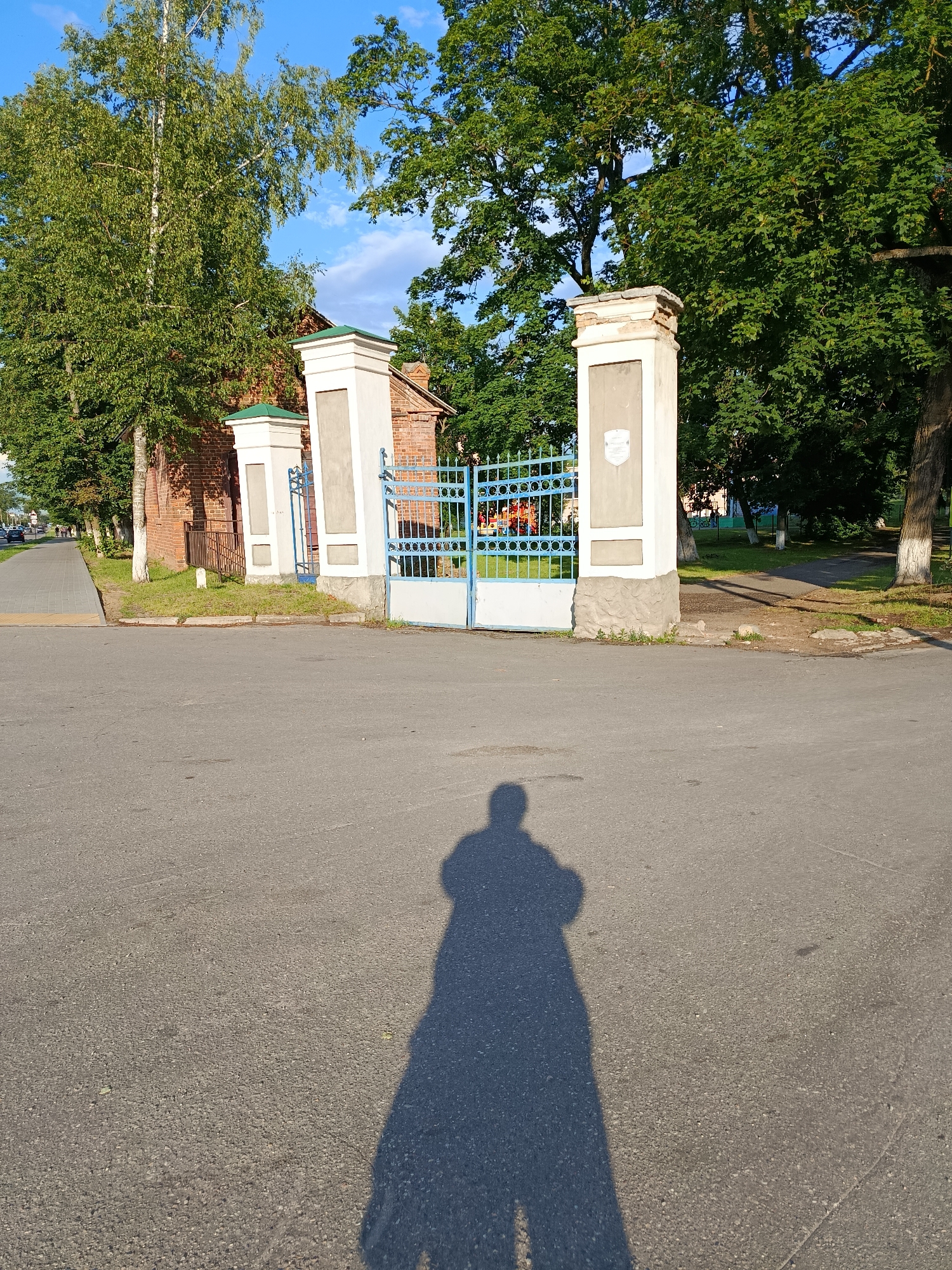 The image depicts an outdoor scene, likely in Smaliavicki Sel'ski Savet, Belarus, with a focus on an entrance to a property. In the foreground, the paved asphalt road occupies the lower half of the frame, with a prominent shadow of a person, presumably the photographer, cast across it. The shadow shows the outline of a person standing.
The midground features a blue metal gate, partially open, providing access to a courtyard or grounds. Flanking the gate are two ornate brick pillars, each topped with white decorative elements and a green roof. The pillar on the left is closer to a red brick building with a visible chimney. The pillar on the right is set against a backdrop of lush green trees.
Beyond the gate, glimpses of what appears to be a playground with colorful structures can be seen. The left side of the image shows a paved pedestrian walkway running parallel to a line of trees and the brick building. A few cars are visible further down the walkway, indicating a road and the presence of traffic. The right side of the midground is dominated by mature trees with dense green foliage, with a pathway receding into the distance. The overall environment suggests a sunny day with clear blue skies dotted with a few clouds.