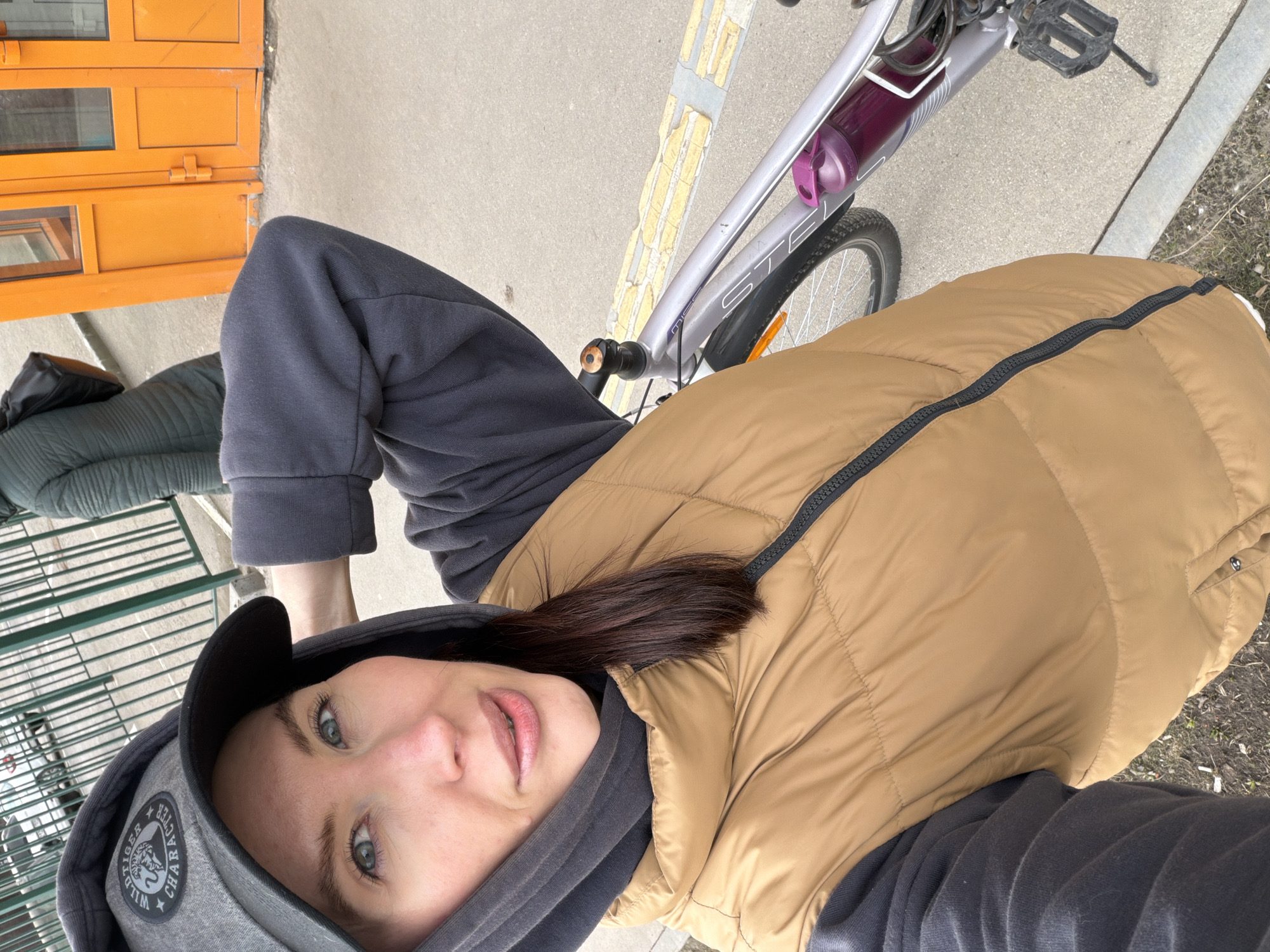 This media file captures a selfie-style portrait of a person (likely female) taken outdoors in Perm, Russia. The individual is looking directly at the camera with a neutral to slightly pleasant expression, showcasing blue-green eyes and dark brown hair peeking from under a grey hooded sweatshirt. She wears a dark grey baseball cap featuring a circular patch with a tiger head design and text, which includes "TIGER FACTOR" and "WILD." Over her hoodie, she wears a camel-colored puffer vest with a black zipper, and dark grey sweatpants are visible on her lower body.
The person appears to be positioned next to a silver bicycle, which is partially visible on the right side of the frame. The bicycle's frame shows the brand "STELS" and has a purple water bottle in its holder. A yellow, peeling decal or sticker is also present on the bike frame. Black pedals and a black fender are discernible.
The background consists of a light-colored paved ground surface. To the left, an orange structure, possibly a kiosk or a small building, features two levels of small, rectangular window panes. A dark bag rests on a green quilted surface in the lower-left background, suggesting outdoor seating or furniture. To the right, sparse vegetation and small weeds are visible along the edge of the paved area. The lighting indicates it is daytime, with diffused light suggesting either an overcast sky or bright ambient light.