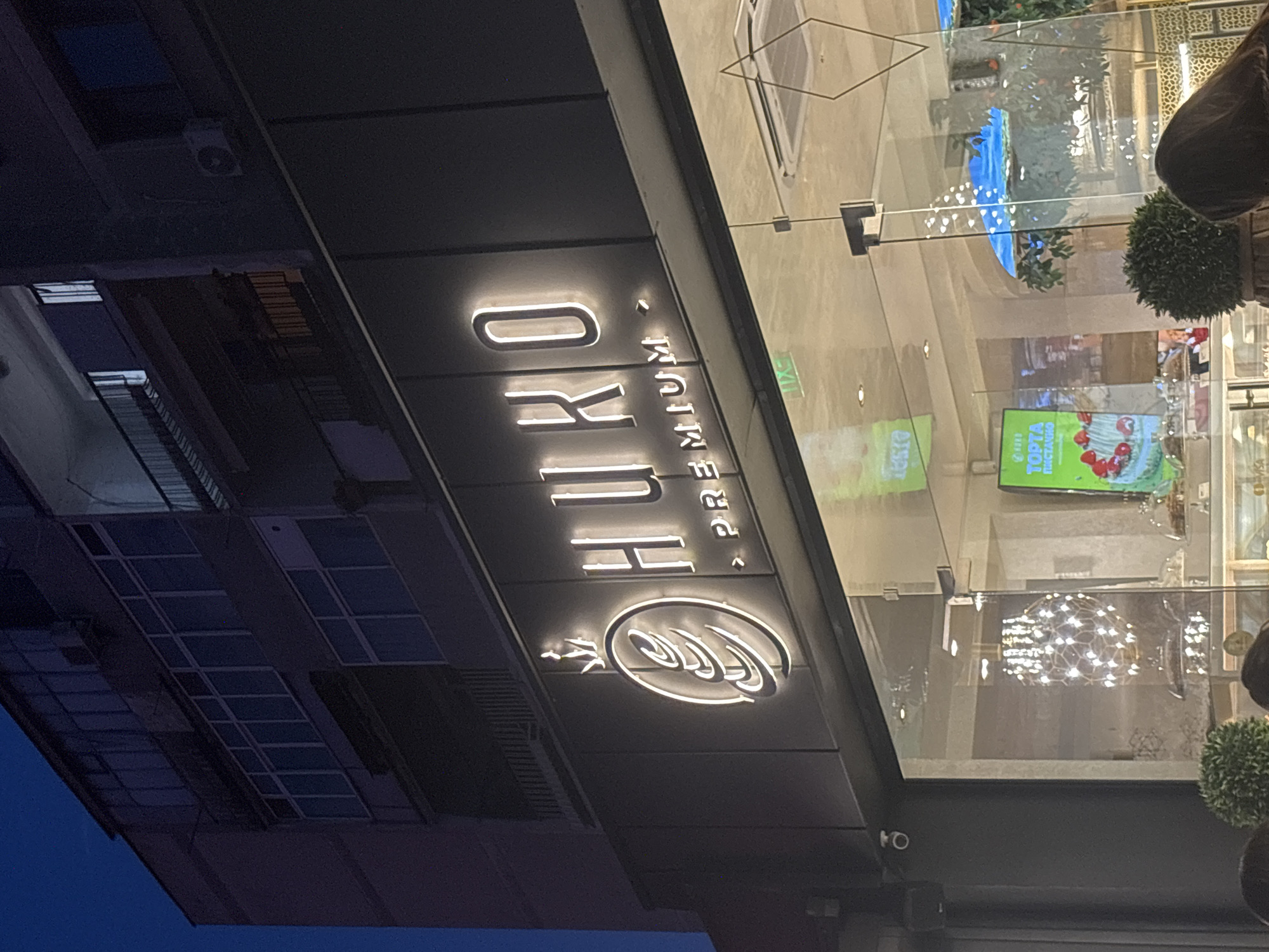 Here's a concise description of the image:
An angled, street-level photograph captures the facade of a shop, identified by a prominent illuminated sign reading "HUKO PREMIUM." The sign features a stylized wheat emblem. The building has a glass-paneled exterior, reflecting interior elements such as lighting fixtures and what appears to be a television screen displaying an image of a cake. The sky is dark. The building is located in Plovdiv, Bulgaria.