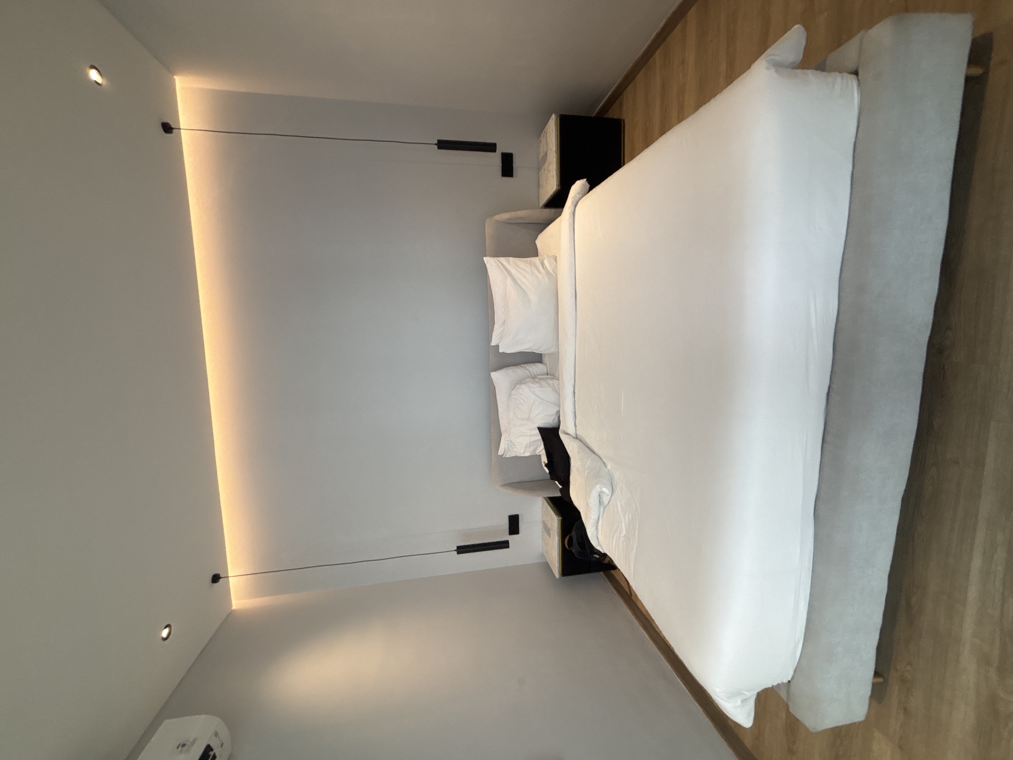 This image depicts a bedroom in North Kuta, Indonesia. The room features a large bed with white linens and several pillows. Two modern bedside tables with marble tops flank the bed, each with a black pendant light hanging above it. The walls are painted a neutral gray, and a strip of warm ambient lighting runs along the top of the wall behind the bed. An air conditioning unit is visible on the upper left wall. The floor is made of light-colored wood. The scene is tidy and suggests a contemporary, minimalist aesthetic.