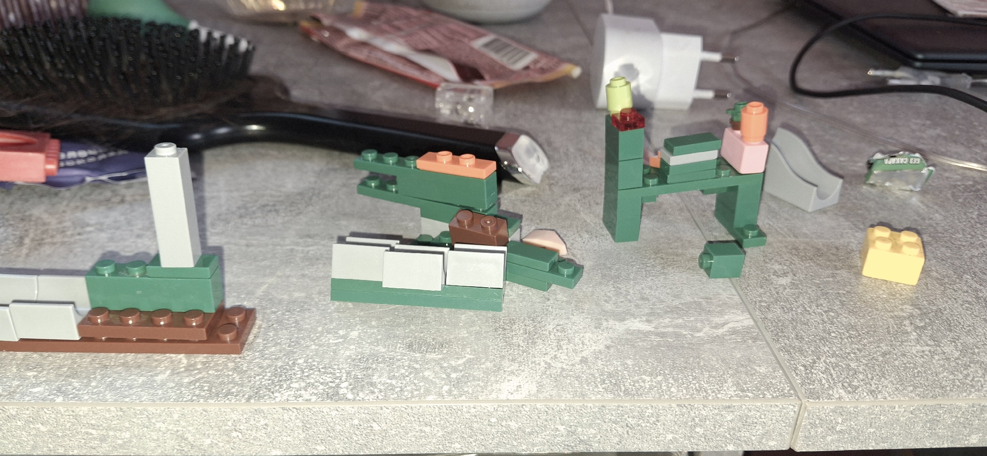 The image depicts a collection of Lego bricks arranged on a gray countertop. The bricks are primarily green, white, brown, and other assorted colors, and are arranged in small, abstract structures resembling animals and other constructions.
Other objects in the scene include a hairbrush, a charging block, packaging for an unknown item, and small electronic components. The setting appears to be a domestic environment.
There is no evidence to suggest this scene is located in Писарёвка, Russia, or any specific location for that matter.