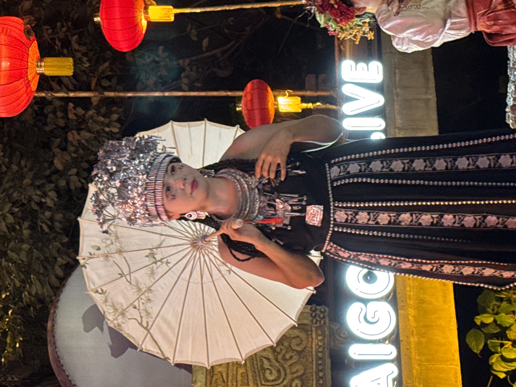 A woman in traditional attire, holding a white umbrella, stands in front of a neon sign that reads "SAIGON.LIVE". She wears a silver headdress, multiple layered necklaces, and a dark dress with embroidered patterns. Her face is adorned with makeup, and she has braids. Red lanterns hang in the background, suggesting an outdoor evening event. The setting appears to be in Yuzhong District, China, potentially near a cultural performance or festival. To her right, a portion of another person in similar festive clothing is visible.