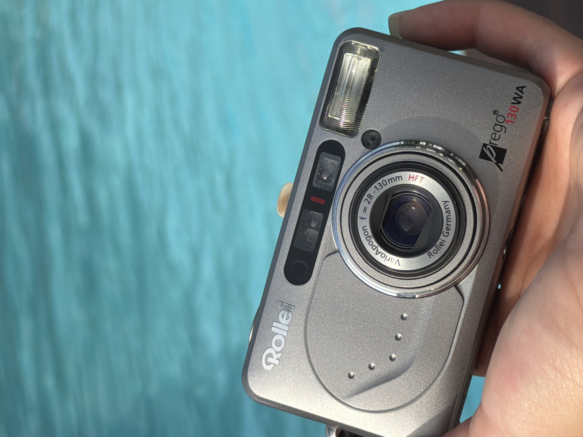 A light-skinned hand holds a silver-grey Rollei prego 130WA compact camera in the foreground, positioned over a bright, rippling light blue body of water. The camera is slightly angled, revealing its front features and branding.
The camera body is a matte silver-grey with "Rollei" embossed on the bottom left. On the top right, "prego®" is printed in black, followed by "130WA" in red text. The lens barrel is prominently featured, displaying the text "VarioApogon f= 28-130mm HFT" and "Rollei Germany." A flash unit is visible on the top left corner of the camera, above a small set of black buttons or indicators. The camera also features raised circular dots on its lower right front, potentially for grip or design.
The background is a uniform expanse of vibrant light blue water, showing subtle wave patterns, which suggests a swimming pool or a calm sea. The bright illumination implies a sunny daytime setting. The camera is held steadily, suggesting it's either about to be used or has just been used. This scene is likely set in Varna, Bulgaria, by a pool or overlooking the Black Sea.