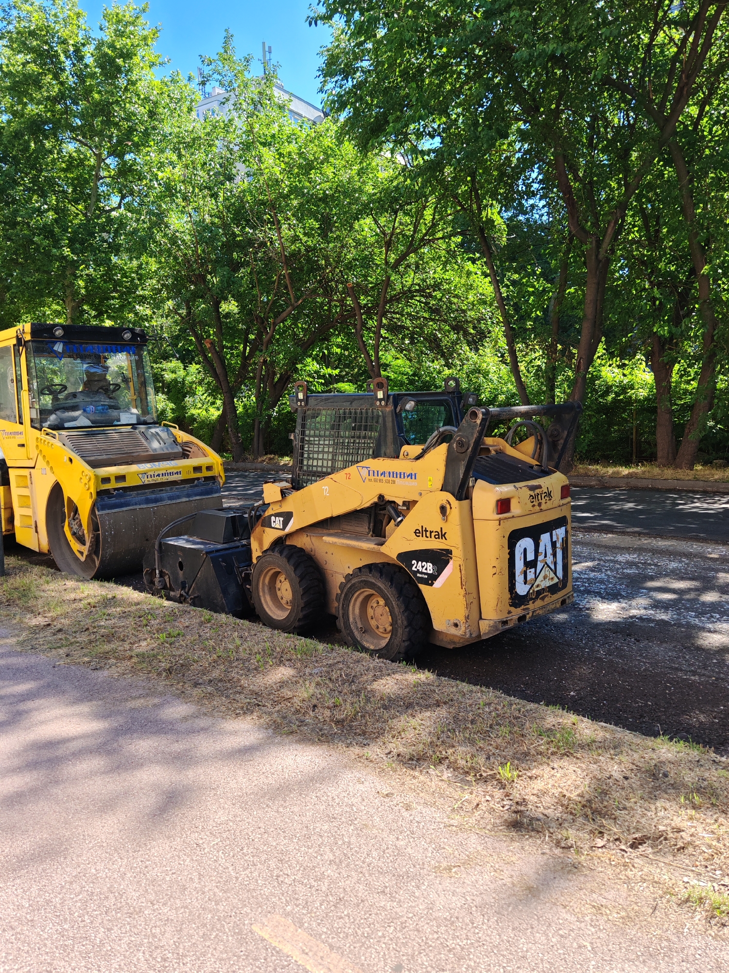 A bright yellow road roller and a yellow Caterpillar skid steer loader are parked on a paved surface next to a grassy area. The road roller, a Bomag brand, has a large cylindrical drum in the front. The skid steer loader, a CAT 242B2 model, is positioned to the right of the roller. The CAT loader has a bucket attachment in the front. Both vehicles appear to be engaged in road construction or maintenance work, as evidenced by the freshly laid asphalt and the presence of construction equipment. The background consists of lush green trees and a clear blue sky, suggesting a sunny day. A white building with multiple antennas on its roof is visible through the trees in the distance. The overall scene captures a moment of urban infrastructure work.