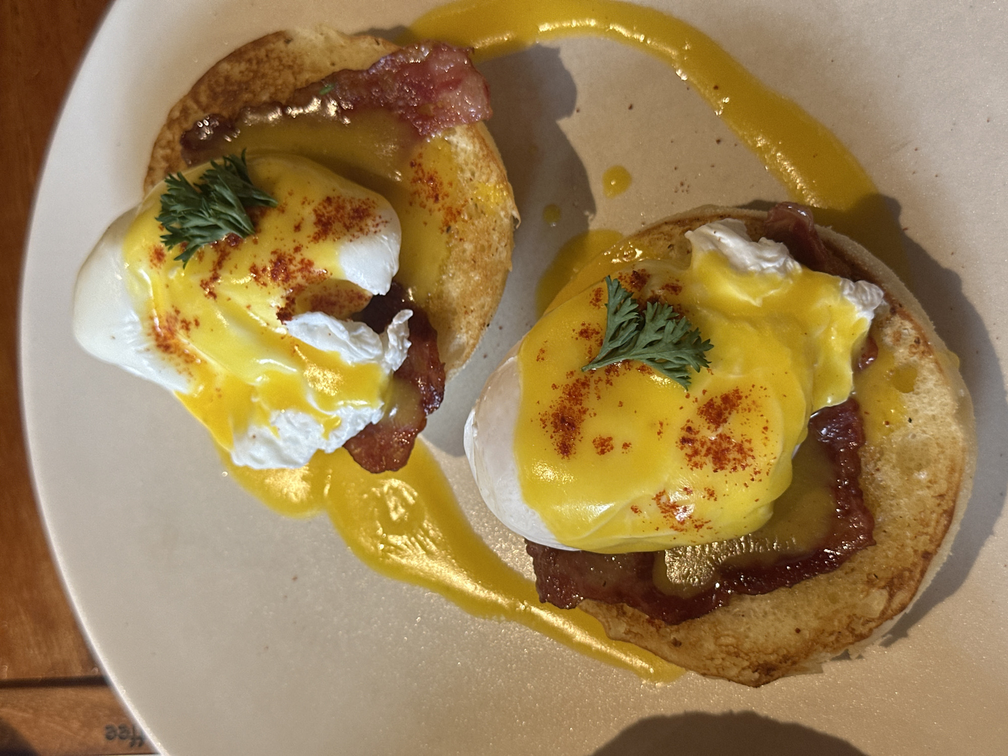 Two servings of Eggs Benedict are presented on a light-colored plate. Each serving consists of a halved English muffin topped with a slice of bacon, a poached egg, and a generous drizzle of hollandaise sauce. The poached eggs have a bright yellow yolk and a soft white exterior, and the hollandaise sauce is a vibrant yellow, pooling around the eggs and on the plate. A light dusting of paprika is visible on top of the hollandaise sauce, and a small sprig of parsley garnishes each serving. The background shows a glimpse of a wooden table surface.