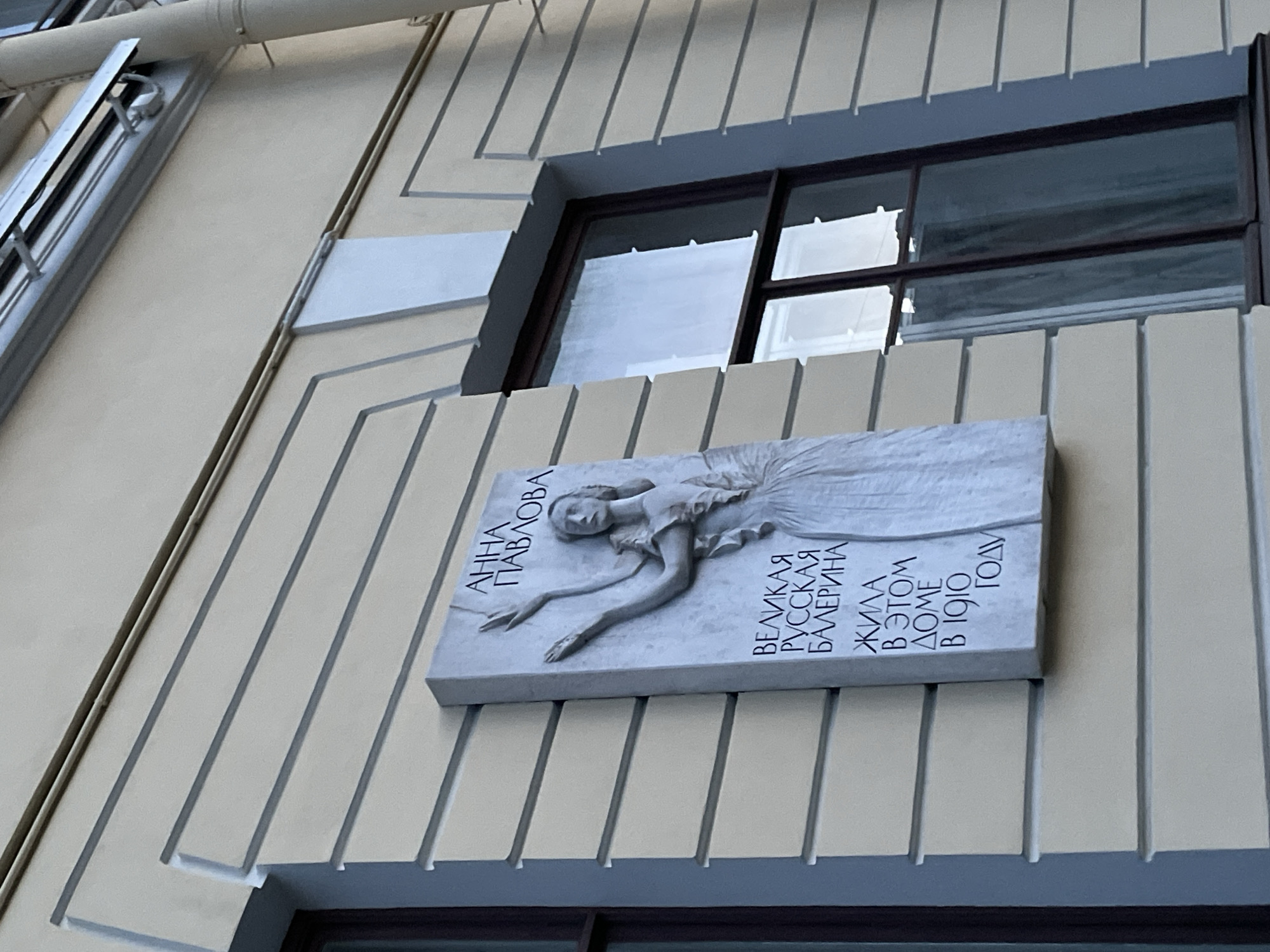 An upward-angled shot captures a light-colored building facade in Saint Petersburg, Russia, featuring a commemorative plaque and a window. The building's exterior is adorned with regularly spaced vertical strips, creating a textured, almost fluted appearance.
Dominating the lower-center of the frame is a rectangular, light-grey commemorative plaque. It displays a bas-relief sculpture of a graceful female figure, presumably a ballerina, lying in an elegant pose with arms extended and wearing a flowing tutu. The details of her face and costume are softly sculpted.
To the left of the sculpted figure, the plaque bears Cyrillic text that reads "АННА ПАВЛОВА" (Anna Pavlova). To the right, further text states: "ВЕЛИКАЯ РУССКАЯ БАЛЕРИНА" (Great Russian Ballerina), followed by "ЖИЛА В ЭТОМ ДОМЕ В 1910 ГОДУ" (Lived in this house in 1910).
Above the plaque, a horizontal rectangular window with dark frames is visible, reflecting a bright, possibly overcast sky or distant buildings. The area immediately surrounding the window is recessed and appears slightly darker than the rest of the facade. On the far left of the image, thin metallic pipes or conduits run vertically up the building. The scene is illuminated by natural daylight, casting subtle shadows and highlighting the relief details of the plaque.