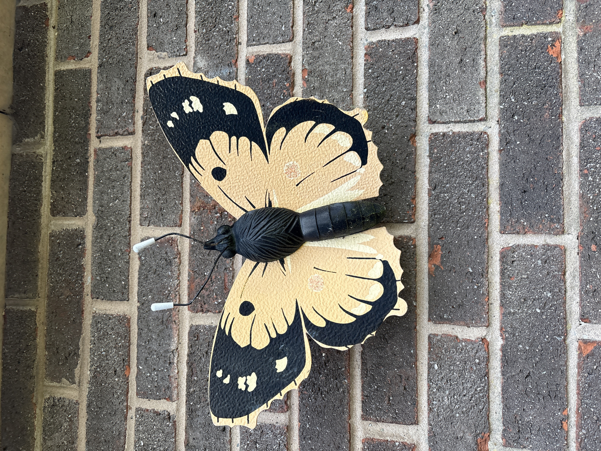 A close-up, eye-level shot reveals a decorative butterfly affixed to a brick wall. The butterfly is predominantly beige with black accents and white-tipped antennae. It is attached to the wall, which is made of dark grey bricks with lighter grout. The scene lacks any people, actions, or interactions. The image provides no information to confirm the location in Minsterley, United Kingdom.