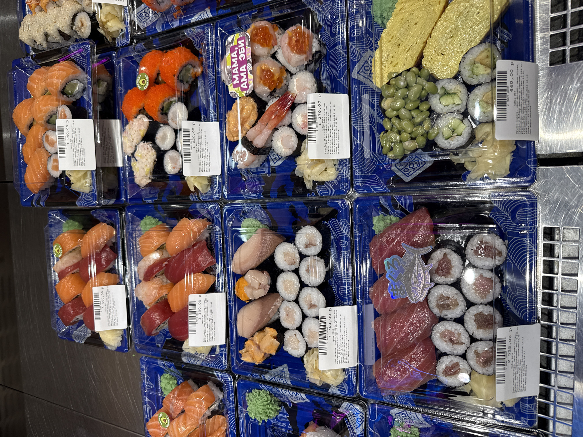 This is a top-down, eye-level view of a display of various pre-packaged sushi sets in a retail environment, likely a grocery store. The setting appears to be indoors, with a metallic shelf structure visible on the right side of the frame, suggesting a cooler or display unit. The lighting is artificial and even, indicating an indoor retail space. The sushi sets are arranged in clear plastic containers, each with a blue and white patterned backing.
The subjects are primarily the sushi sets themselves, which include a variety of rolls and nigiri featuring salmon, tuna, and other seafood, as well as some vegetable-based rolls. Some sets also include edamame, tamagoyaki (rolled omelet), and pickled ginger. The visible text on the packaging includes price information (e.g., "1 100,00 p", "1 020,00 P", "1 740,00 P", "460,00 P", "2 525,00 P", "1 270,00 P"), product descriptions in Russian (e.g., "Набор у", "Набор 324", "Набор 187", "Набор 847"), and production/expiration dates (e.g., "26.06.2024", "26.06.2026"). One container also features a prominent purple label with "MAMA, АМА ЭБИ" in white lettering.
There are no people visible in the image, and the activity is the presentation of goods for sale. The time of day is not specified, but the consistent lighting suggests it could be any time during operating hours. The overall impression is one of organized food display, showcasing a selection of ready-to-eat sushi options. The location context provided is Khimki, Russia, which aligns with the Cyrillic text on the labels.