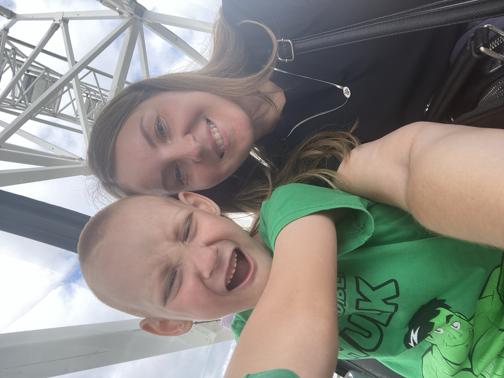A woman and a young boy are taking a selfie in a Ferris wheel cabin. The woman is on the right, smiling, with blonde hair and a black shirt. She is wearing a silver necklace. The boy is on the left, making a funny face, with a green t-shirt featuring the Hulk character. The background shows the white structure of the Ferris wheel and a cloudy sky. The location is the "Izhevsk Eye" Ferris wheel in Izhevsk, Russia.