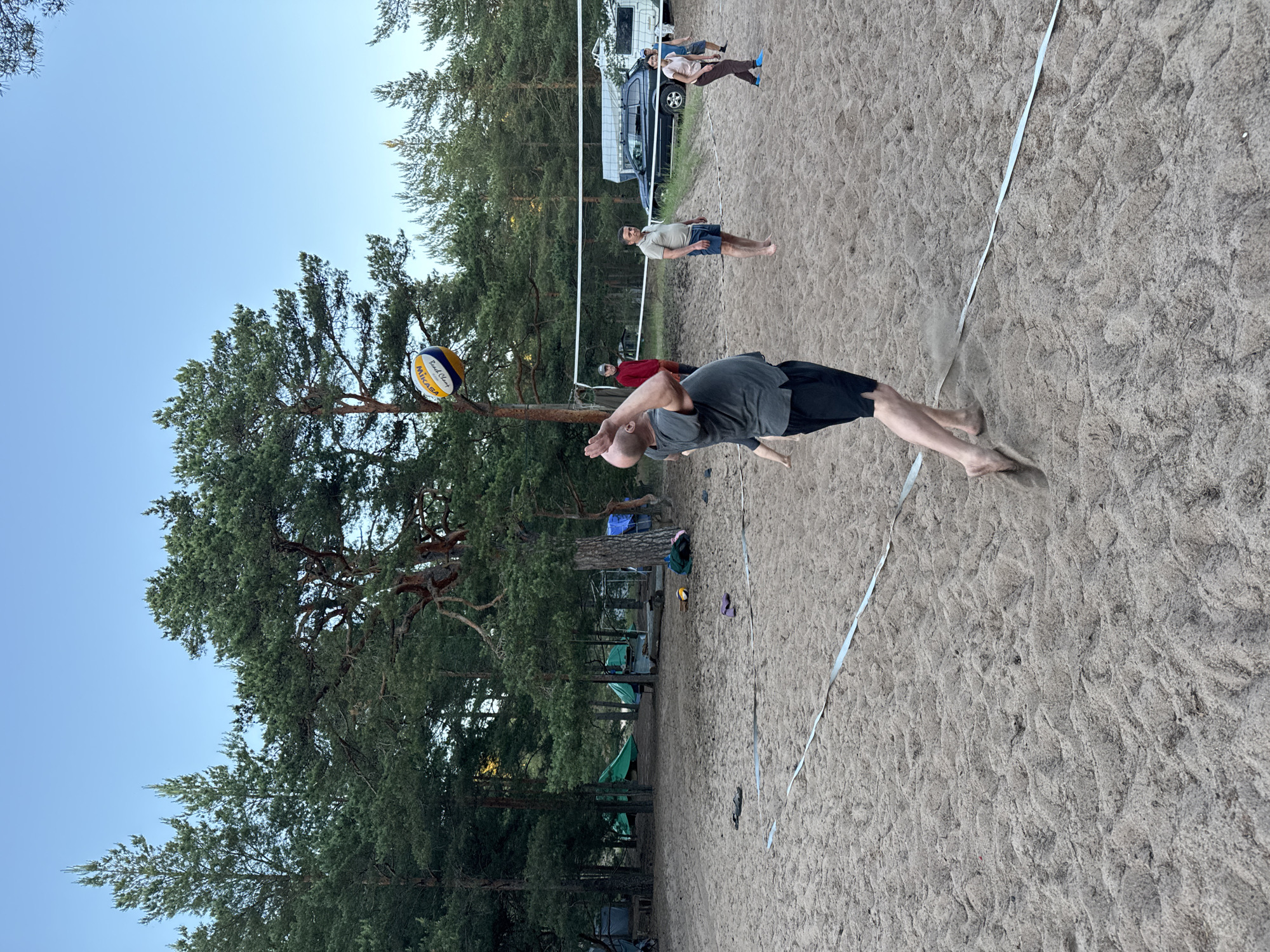 The image depicts a beach volleyball game in Zelenaya Roshcha, Russia. A man in a grey t-shirt and black shorts is in mid-air, serving a Mikasa volleyball. Several other people are visible on the sandy court, including a man in a red shirt, a man in a beige shirt and blue shorts, and a woman in a pink t-shirt and dark pants. In the background, pine trees line the beach, and tents and camping equipment suggest a campground. A car and a white camper van are parked near the volleyball net. The sky is a clear, pale blue, indicating it's likely late afternoon or early evening. The sandy terrain shows the ruts and patterns typical of a beach volleyball court.