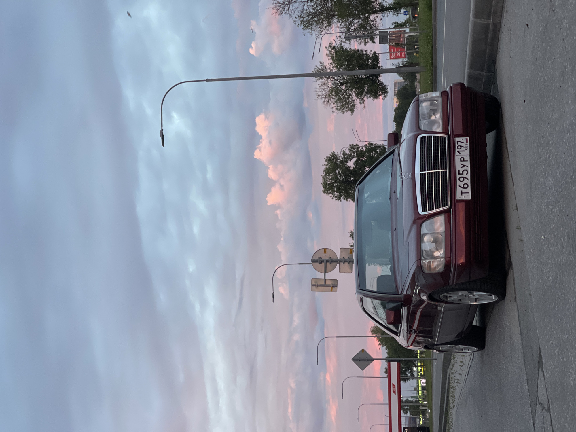 A maroon Mercedes-Benz coupe is parked at the side of a paved road under a cloudy sky with pink and purple hues. The car is angled slightly towards the camera, with its front bumper and grille clearly visible. A license plate with the characters "T695YP 197 RUS" is affixed to the front. Several streetlights and traffic signs are present in the background, along with trees and buildings. A gas station is visible to the left of the car. The overall setting appears to be an urban or suburban environment during dusk.