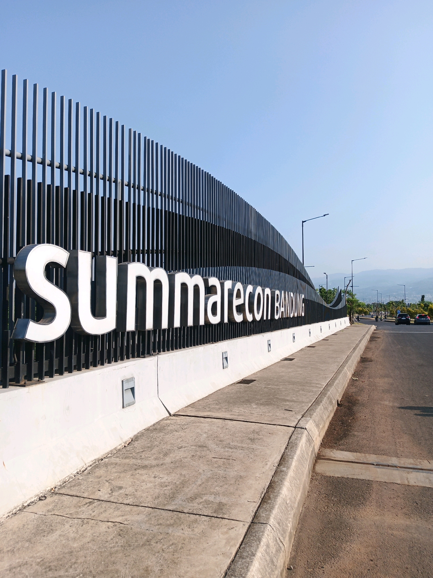 An outdoor, eye-level, medium shot captures the entrance to Summarecon Bandung in Bandung City, Indonesia, under a clear blue sky. The prominent feature is a large, white, three-dimensional sign that reads "SUMMARECON BANDUNG," mounted on a dark, vertical-slatted fence. The fence curves along the side of the road, with a concrete sidewalk and curb running parallel to it. Beyond the fence, a road stretches into the distance, with a few cars visible and mountains in the background. The scene is well-lit by natural sunlight, casting subtle shadows. The architecture of the entrance suggests a modern and well-maintained urban development.