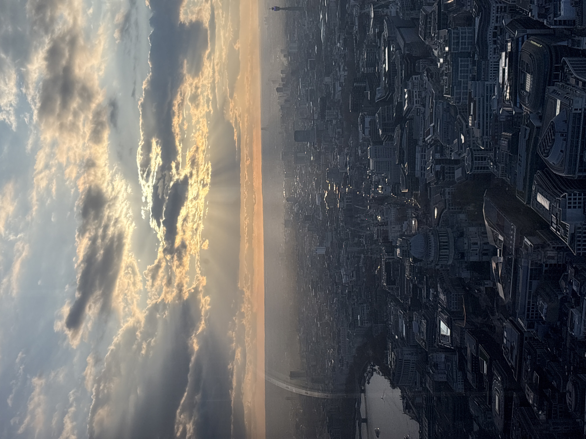 This image shows an aerial view of London, United Kingdom. On the left, the sky is visible with a mix of clouds and sunlight. The right side of the image shows the city skyline with various buildings, including identifiable structures such as the London Eye and the Shard in the distance. The Thames River winds through the city, with a few boats visible on the water. The city is rendered in a monochromatic tone, contrasting with the colors in the sky. The perspective is an elevated angle, suggesting the photo was taken from a high vantage point.