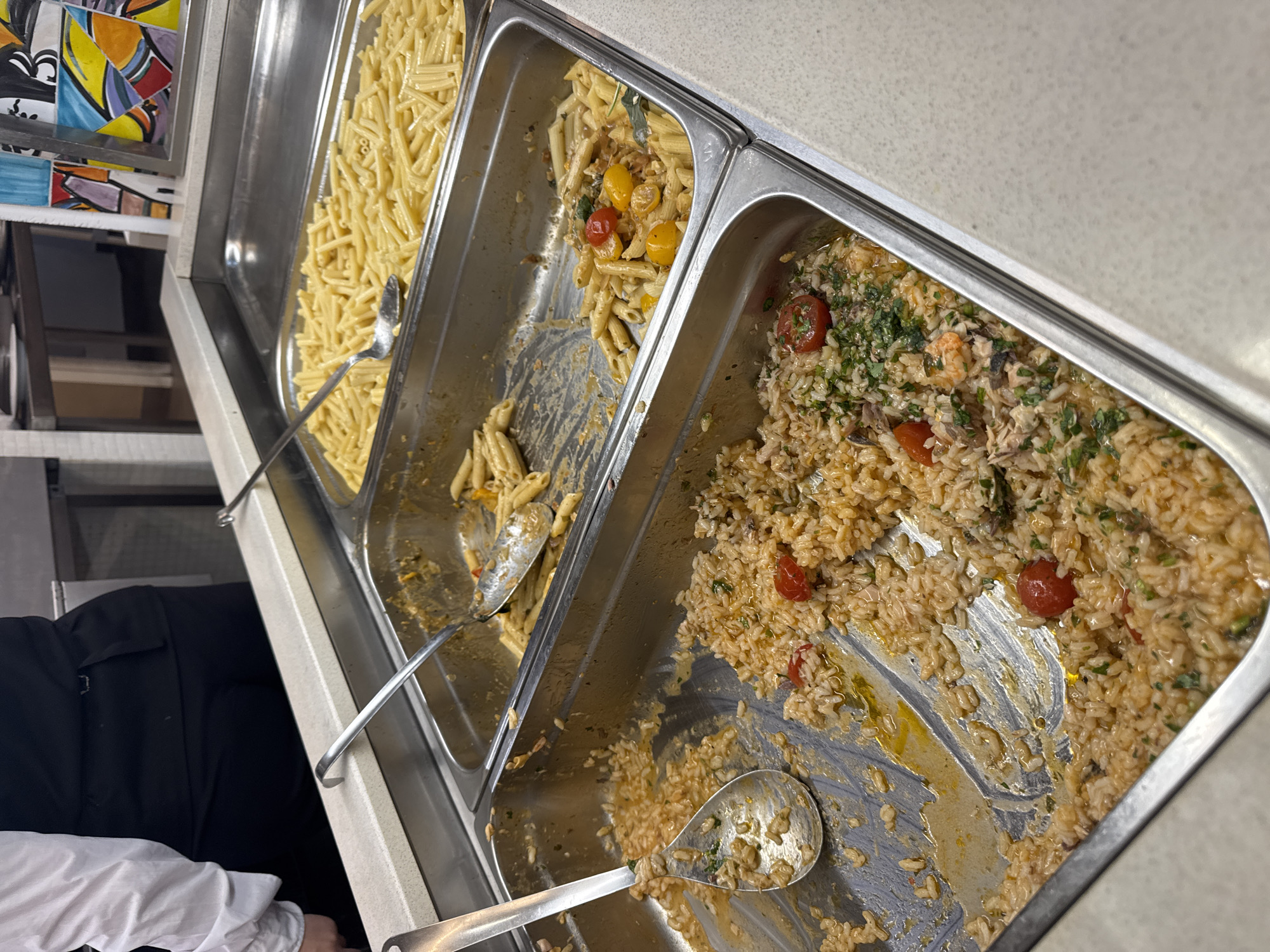 The image shows a buffet line at a restaurant. There are three stainless steel trays filled with food. The tray on the right contains macaroni. The middle tray contains penne pasta with tomatoes and what appears to be bell peppers and other vegetables. The tray on the left contains rice with shrimp, tomatoes, and herbs. A person in a black vest and white shirt is partially visible in the background. The restaurant appears to be located in Isola delle Femmine, Italy.