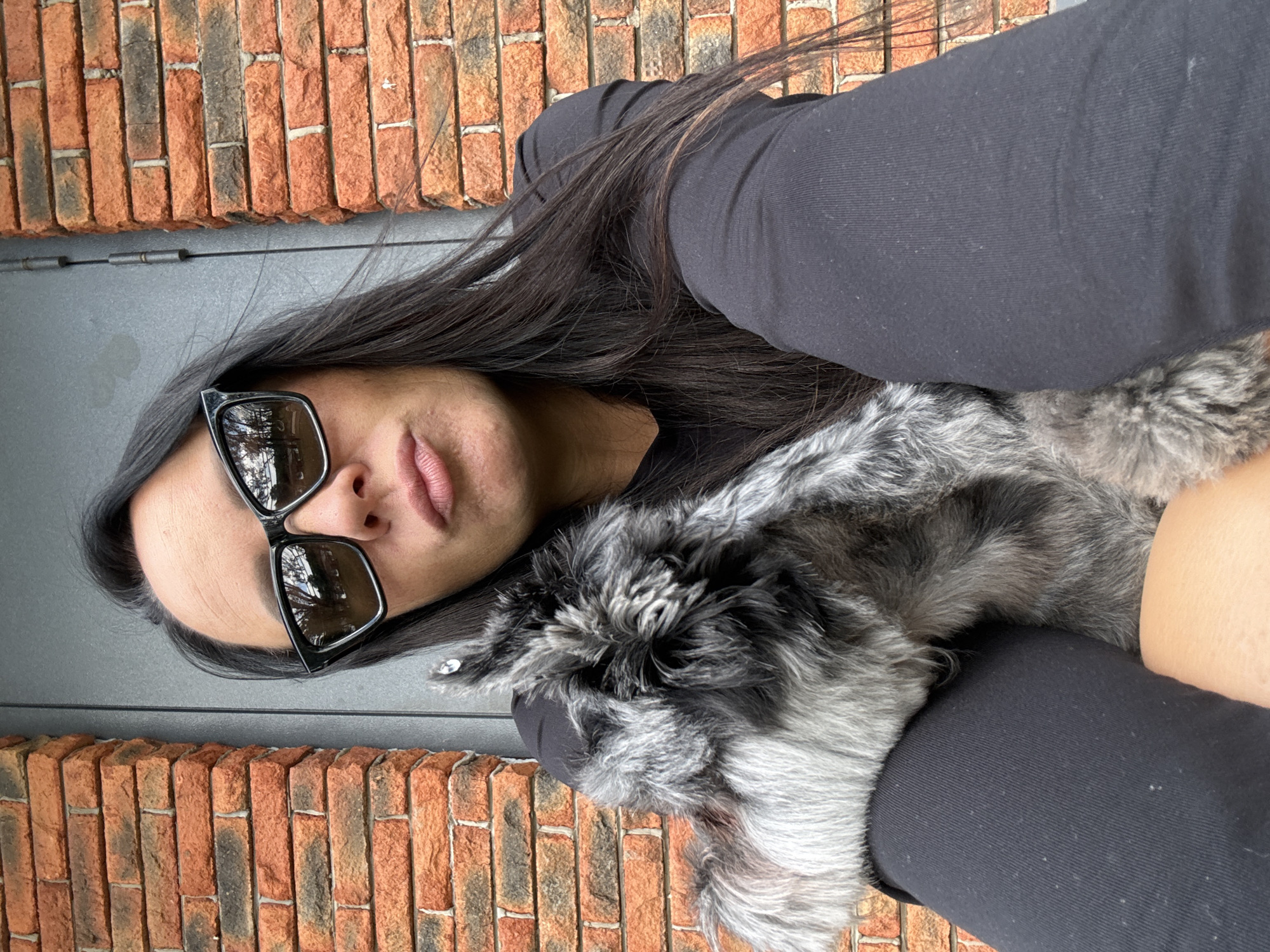 A medium shot captures a woman from the chest up, outdoors, embracing a small, shaggy dog. The woman, wearing black leggings, a black shirt, and black-rimmed sunglasses, has dark hair that falls past her shoulders. Her right arm is around the dog, which is predominantly grey with patches of black and white fur. The dog is nestled against her left shoulder. The background features a brick wall on the left and a dark grey door with a vertical seam on the right, suggesting an urban environment, possibly a residential building. The lighting is neutral, indicating daytime.