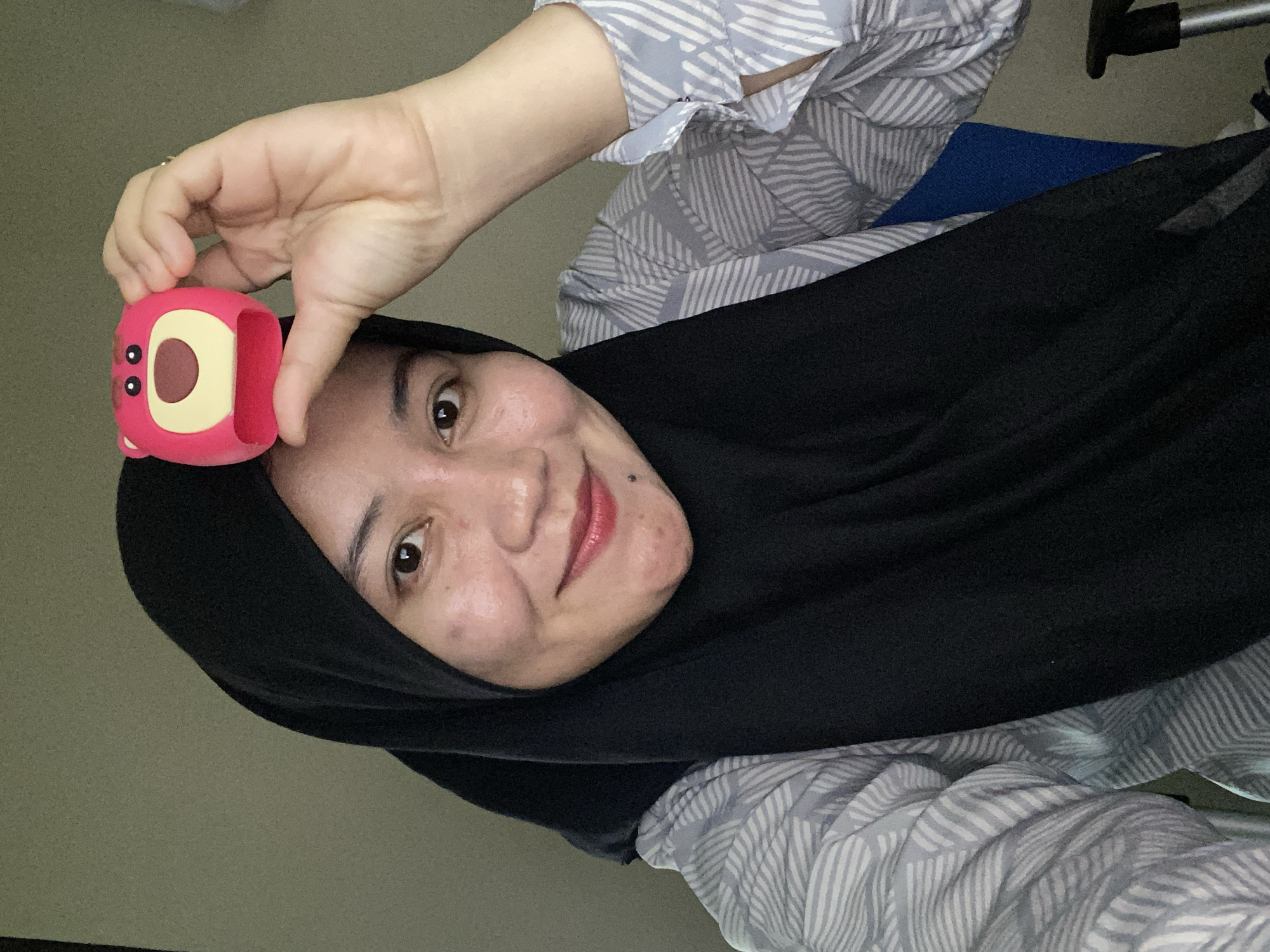 A medium shot captures a woman wearing a black hijab and a light blue and white striped collared shirt. She is looking at the camera and smiling, with her right hand raised to the top of her head, holding a small, pink, bear-shaped object. The background is a plain, light brown wall. The scene appears to be indoors, possibly in a home or office environment. The woman's facial features are visible, including her brown eyes, red lipstick, and a mole on her chin. There are some minor skin blemishes visible on her cheeks. The lighting is diffused and appears to be coming from the front. The overall impression is a casual, informal selfie. Banjarbaru, Indonesia is mentioned, but there are no specific geographical markers within the image to confirm this location.