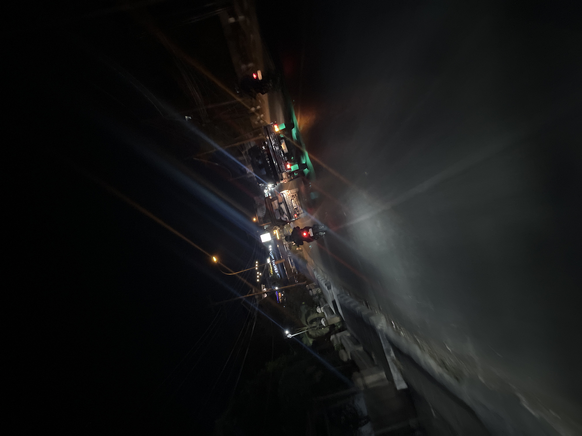 The image captures a nighttime street scene in Kalinyamatan, Indonesia. The perspective suggests the photographer is on a moving vehicle, possibly a motorcycle, looking forward. The road surface is visible, appearing dark and wet, reflecting the lights of the vehicles.
Several vehicles are present. In the foreground, a motorcycle is being ridden, with its taillight illuminated. Behind it, two pickup trucks are driving, both with their brake lights and some neon green undercarriage lighting illuminated. The truck further ahead has a license plate that reads "G 8309 XZ".
The environment includes a sidewalk on the left side of the road, lined with low walls and some greenery, including palm trees. Streetlights are visible, casting a warm glow. Buildings with illuminated signs are present in the background, suggesting a commercial or residential area. Power lines crisscross overhead. The overall impression is a moderately busy urban street at night, with the characteristic lighting and atmosphere of a Southeast Asian city.