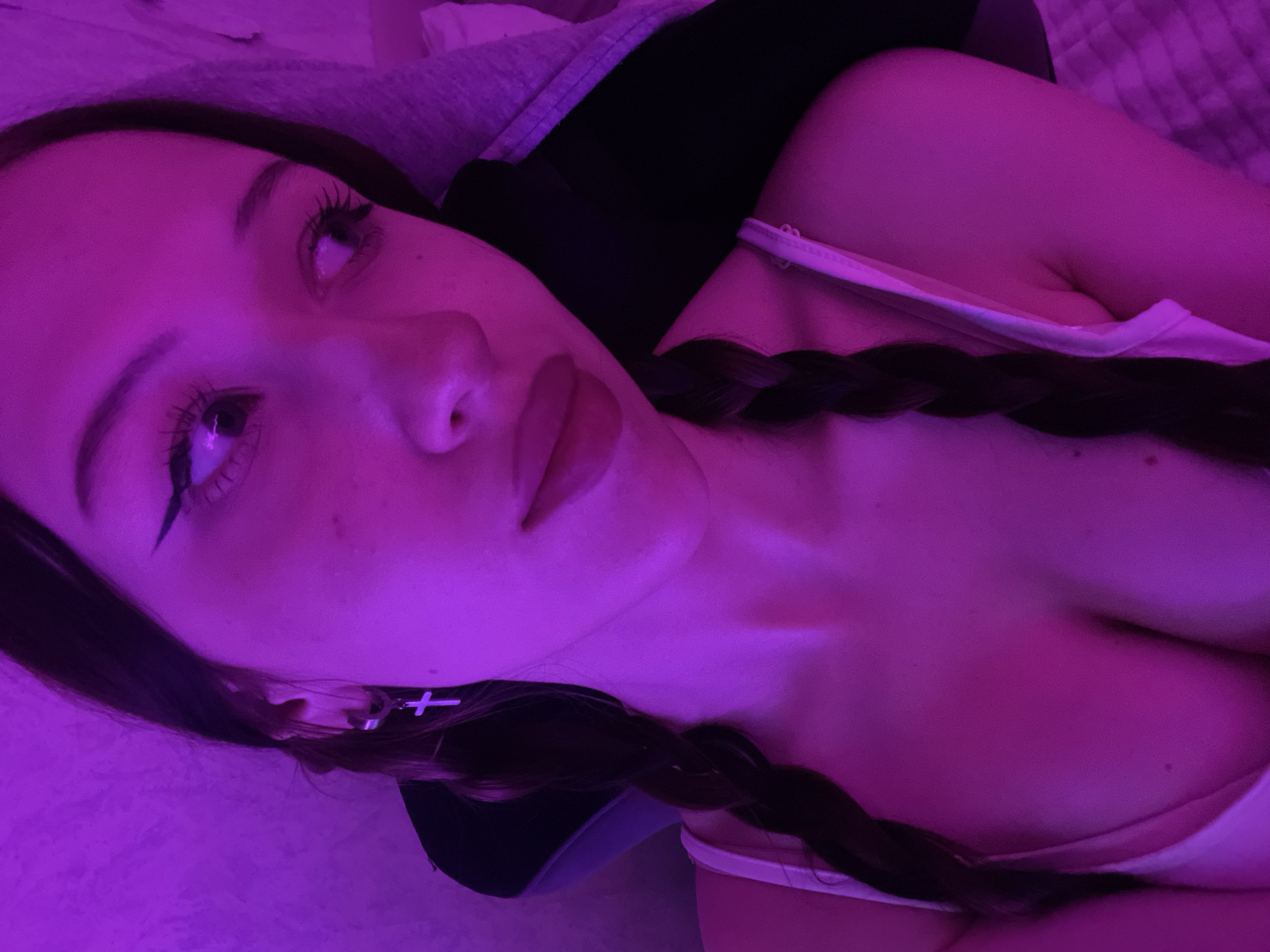A close-up, high-angle portrait features a young person, likely female, illuminated by strong magenta-purple lighting. The subject has dark hair styled in two visible braids, one draped over each shoulder. Eyeliner with a prominent wing, mascara, and defined eyebrows are visible. A silver-toned hoop earring with a dangling cross charm is worn on the visible right ear. Slight freckles are present on the cheek. The person's lips are full and appear to have lipstick applied. She is wearing a light-colored, possibly white or cream, tank top or camisole with thin shoulder straps, revealing a partial view of her upper chest. A dark garment, possibly a hoodie, is partially visible behind her shoulders. The subject's gaze is directed upwards and to the right, away from the camera. The background is indistinct, suggesting an indoor setting with a textured surface on the left and a darker, possibly upholstered, area on the right.