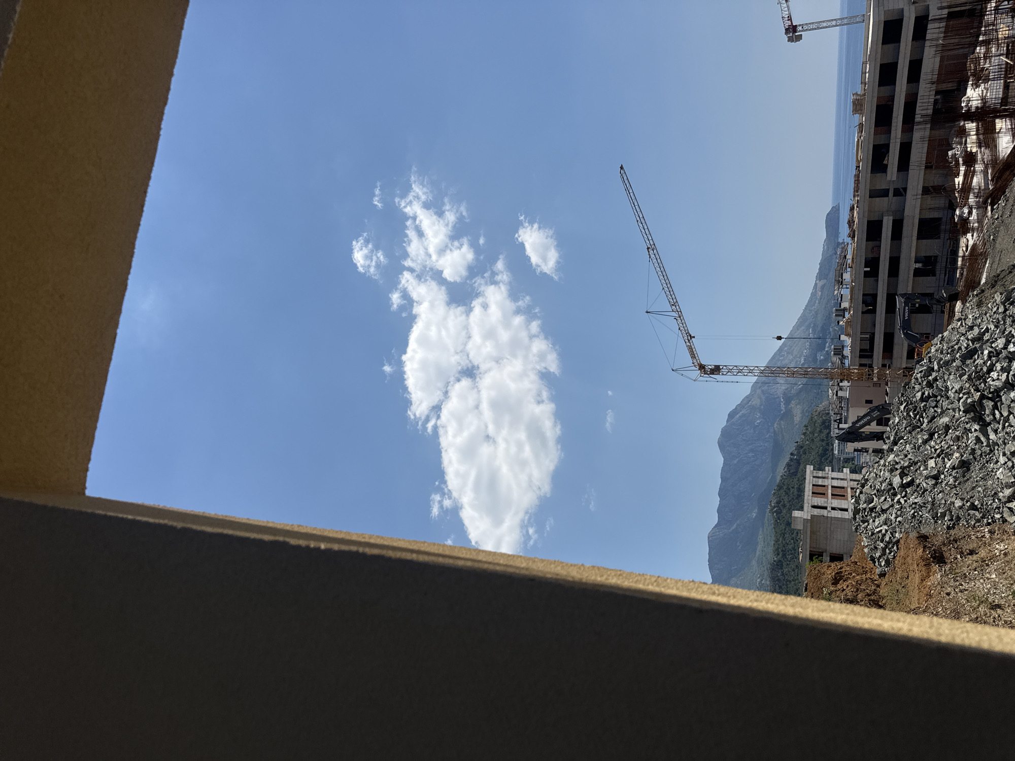 An outdoor, eye-level shot captures a construction site in Boreti, Montenegro, on a clear, sunny day. The foreground is dominated by a large, rough-textured, beige-colored architectural element, likely part of a building under construction, that frames the scene from the left. Beyond this, a vast expanse of bright blue sky stretches across the upper portion of the image, dotted with a few white, fluffy clouds.
In the middle ground, the construction site itself is evident. A prominent yellow tower crane extends across the sky, its jib angled towards the right. Below the crane, a multi-story building is in various stages of completion, characterized by its concrete structure and dark, open window frames. Piles of gray rubble and construction materials are scattered across the ground. To the right of the main building, another, smaller tower crane is partially visible against the horizon.
The background reveals a picturesque landscape. Rolling green hills rise up, transitioning into rugged, dark mountains in the distance. The coastline and the Adriatic Sea are visible on the right side of the frame, with the gentle curve of the sea meeting the land. The overall impression is one of ongoing development against a scenic natural backdrop. There are no visible people in the image.