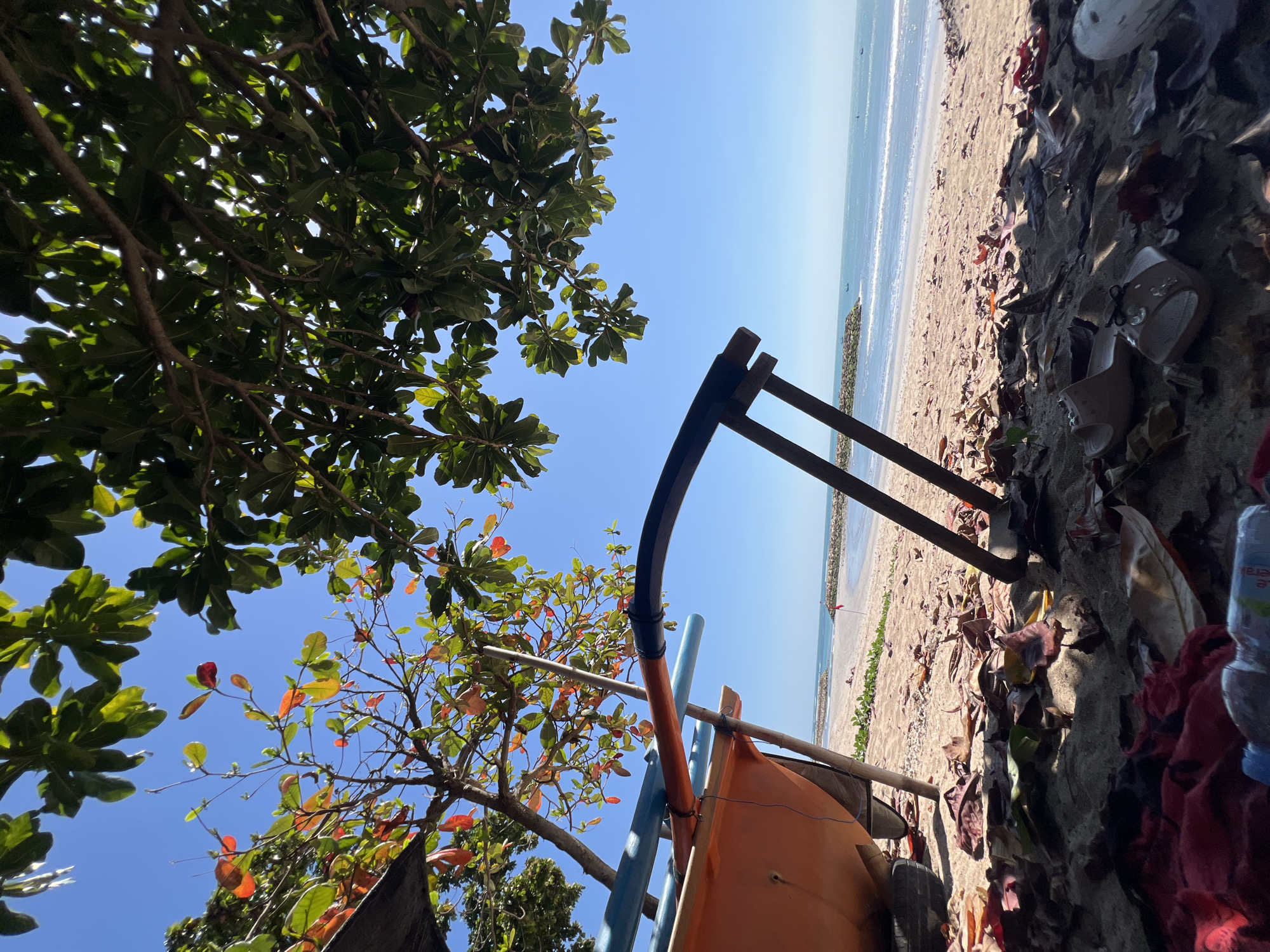 An eye-level, wide shot captures a tranquil beach scene in Kuta, Indonesia, on a bright, sunny day. The foreground is dominated by the sandy beach scattered with fallen leaves and the base of an orange boat. The boat is propped up by wooden beams and has blue and orange poles extending from it. Two wooden posts support a dark curved object, possibly part of a canopy. In the mid-ground, the calm ocean stretches out towards a rocky breakwater with a red flag planted on it. A few small boats can be seen in the distance. Lush green trees with some reddish-orange leaves provide shade overhead, with their branches and foliage framing the clear blue sky. The scene is devoid of any visible people.