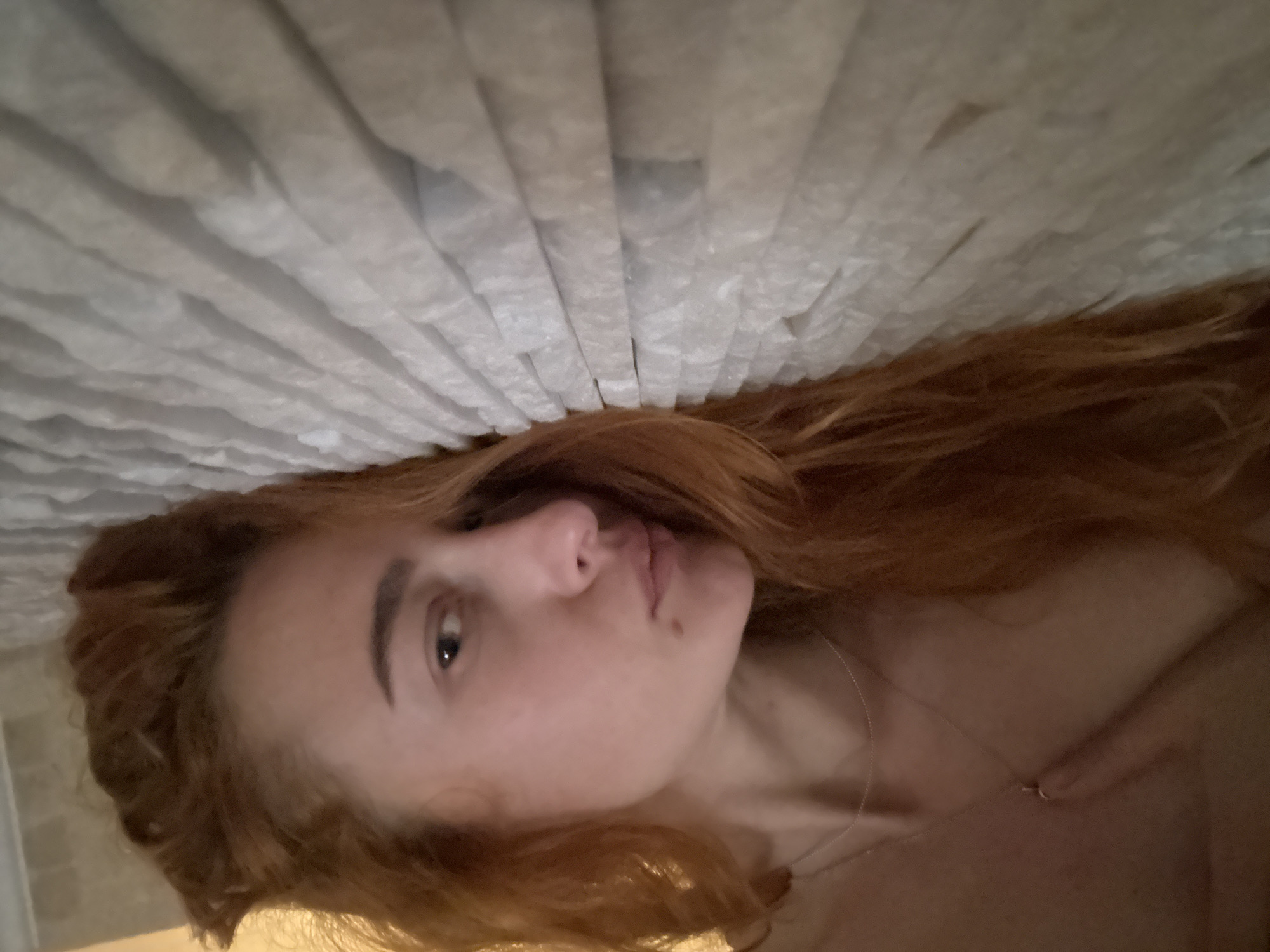 The image shows a woman with long, red hair. She is lying down, with her head near a textured, white surface composed of repeating vertical lines, possibly a headboard. The camera angle is from above, looking down at her face. She has fair skin, light eyebrows, and a mole near her lips. A delicate necklace is visible on her chest. The image provides no information about the location in Budva, Montenegro.