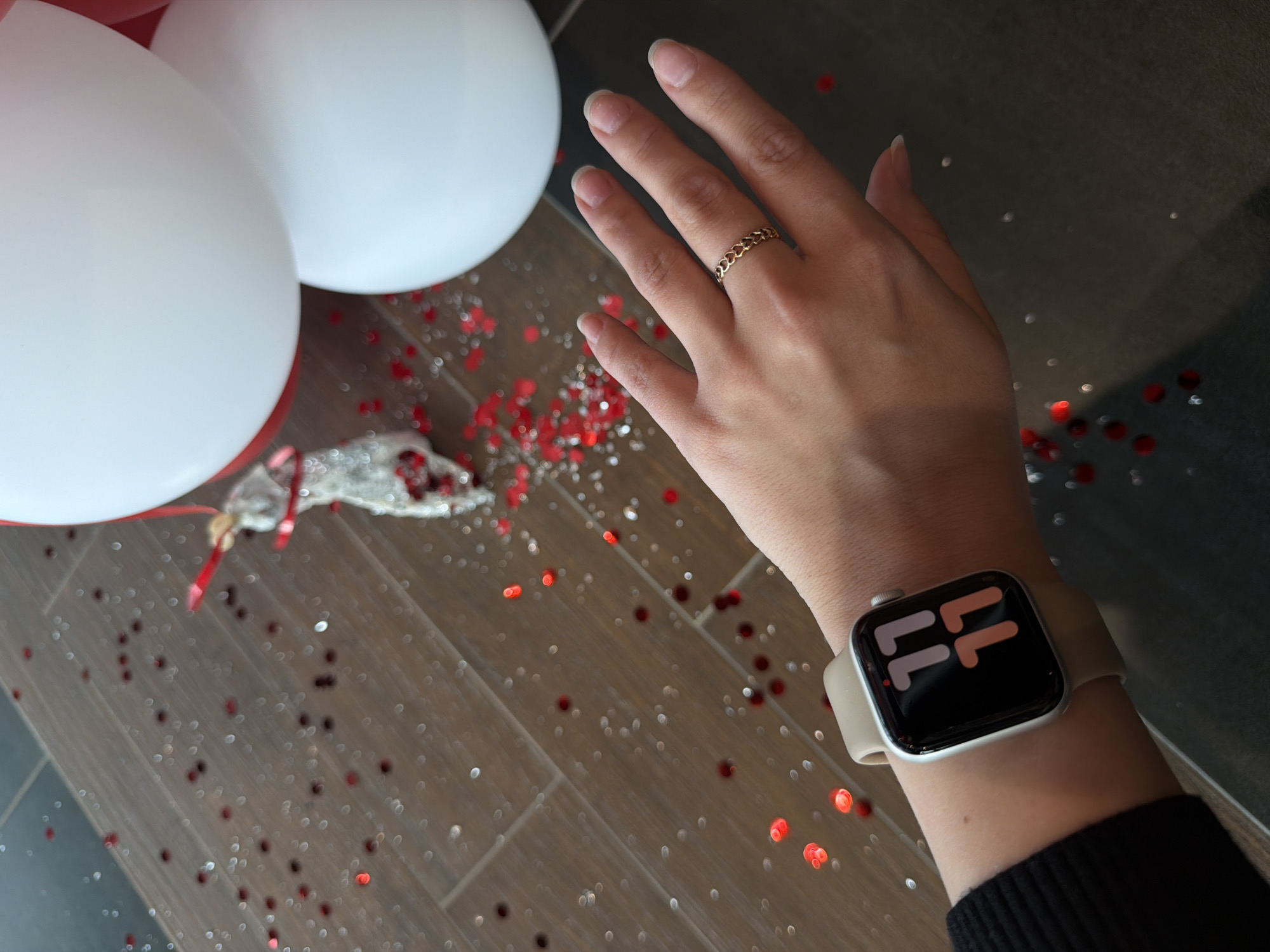 A hand with a gold ring is extended over a scene featuring white and red balloons, confetti, and an Apple Watch on the wrist. The background is a tiled floor. The image does not contain sufficient information to confirm the location in Izhevsk, Russia.
